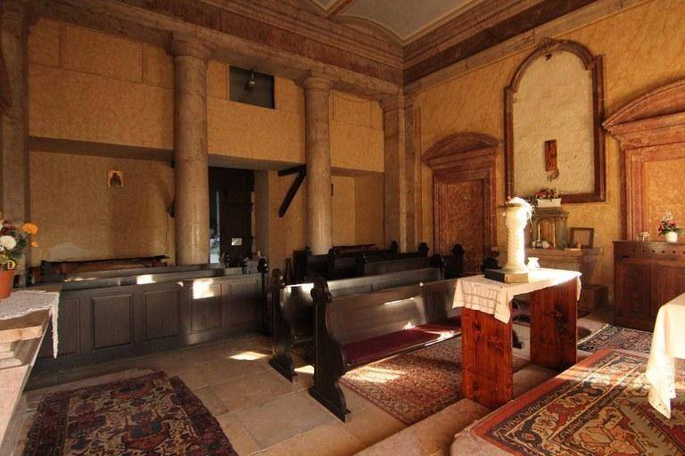 Hellerer Blick in eine historische Kapelle mit Säulen, Holzbänken, gemusterten Teppichen und einer Nische.