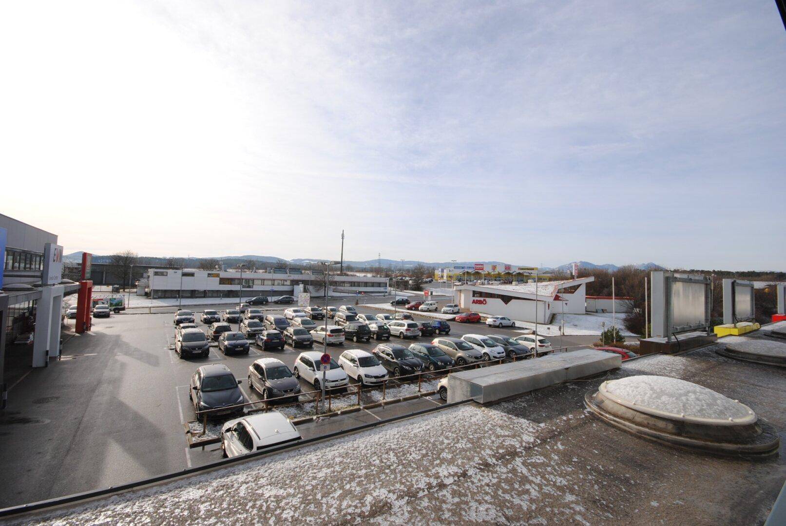 Weitläufiger Blick über einen großen Parkplatz und umliegende Geschäftsgebäude mit Bergen im Hintergrund.