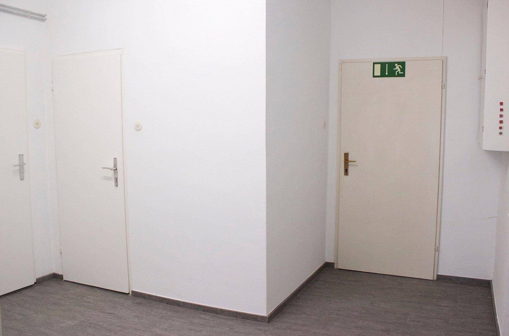 Schlichter Flur mit zwei weißen Türen, eine davon als Notausgang gekennzeichnet.