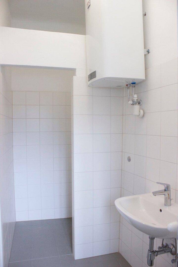 Badezimmer mit Duschbereich, Waschbecken und einem an der Wand montierten Warmwasserbereiter.