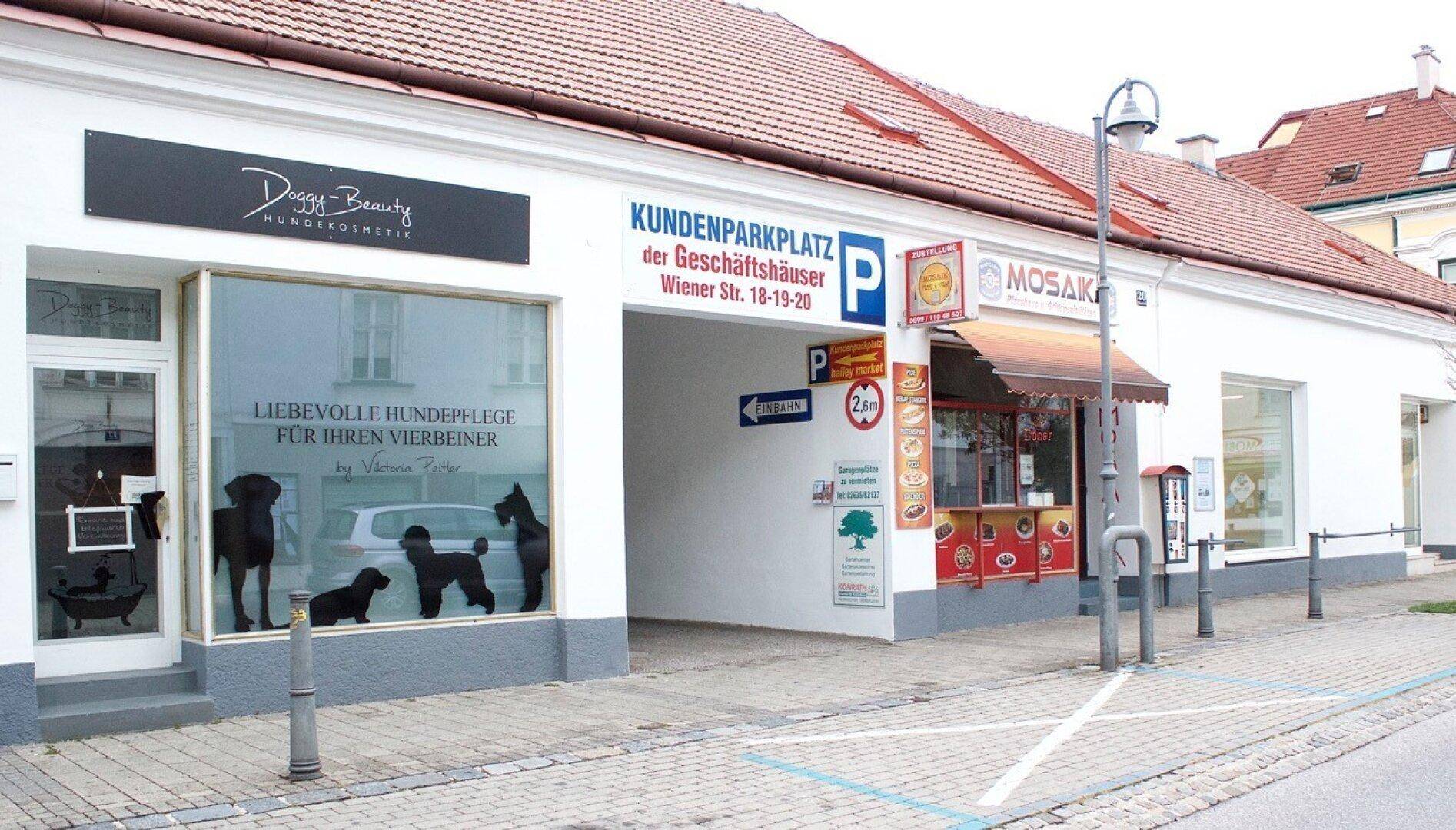 Geschäftsgebäude mit Einfahrt zum Kundenparkplatz und verschiedenen Ladenfronten an der Straße.