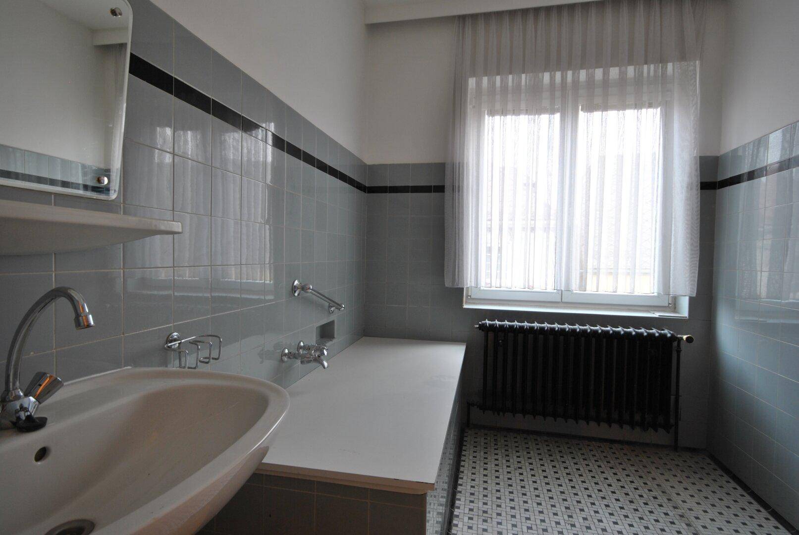 Badezimmer mit grauen Fliesen, Waschbecken und einer integrierten Badewanne, ausgestattet mit Haltegriffen.