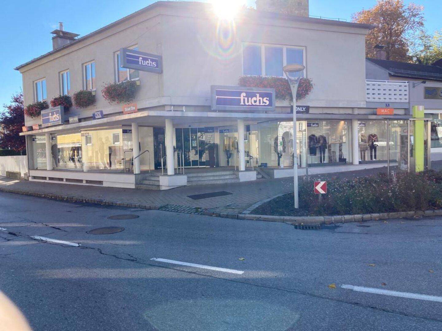 Geschäftshaus an einer Straßenecke mit großen Schaufenstern und Werbeschildern unter strahlendem Sonnenschein.