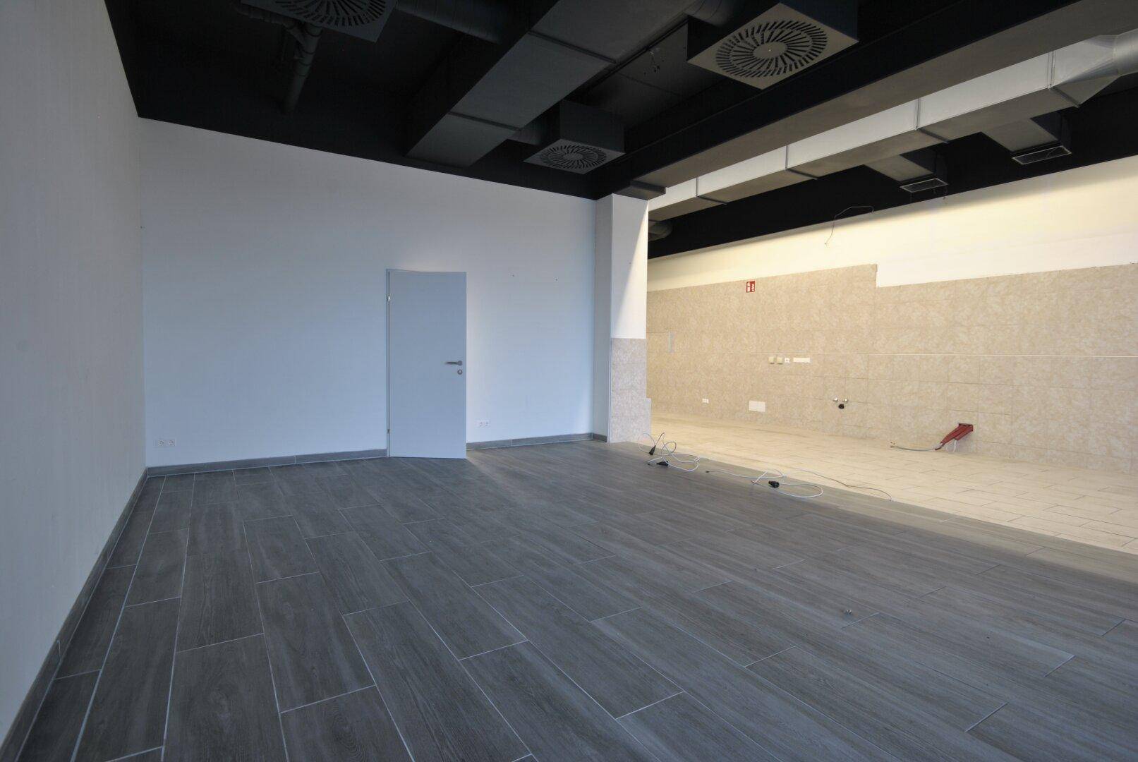 Heller Gewerberaum mit dunklem Holzplankenboden und einer modernen schwarzen Decke mit sichtbaren Lüftungselementen.