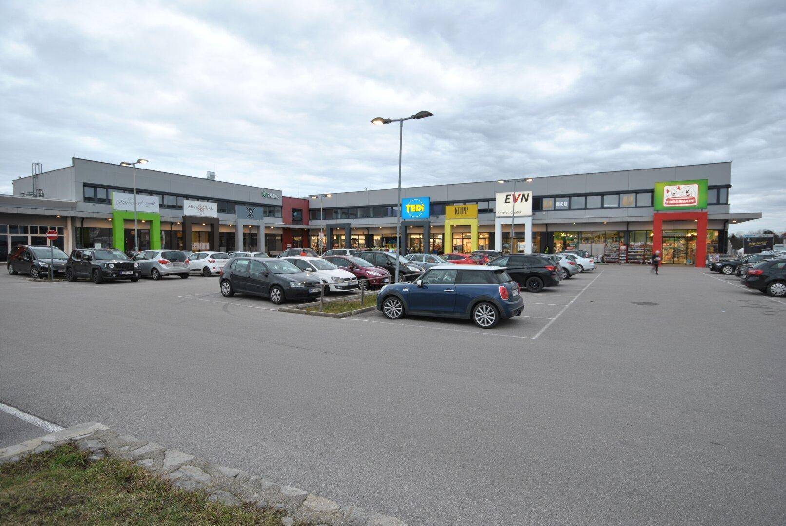Weitläufiger Gewerbekomplex mit großem Parkplatz und verschiedenen Geschäften unter bewölktem Himmel.
