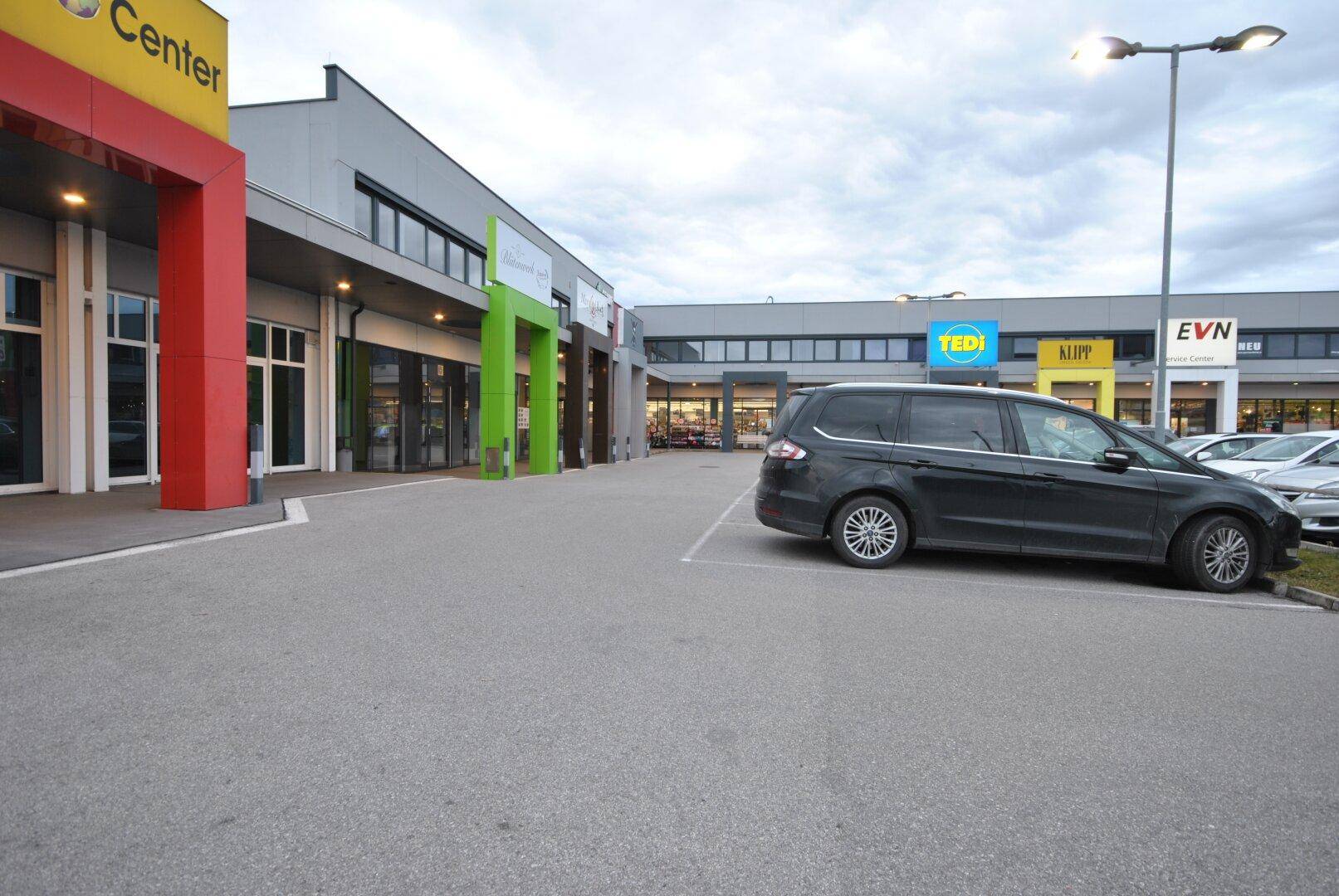 Außenansicht eines modernen Geschäftszentrums mit verschiedenen Ladenfronten und einem großen Parkplatz.