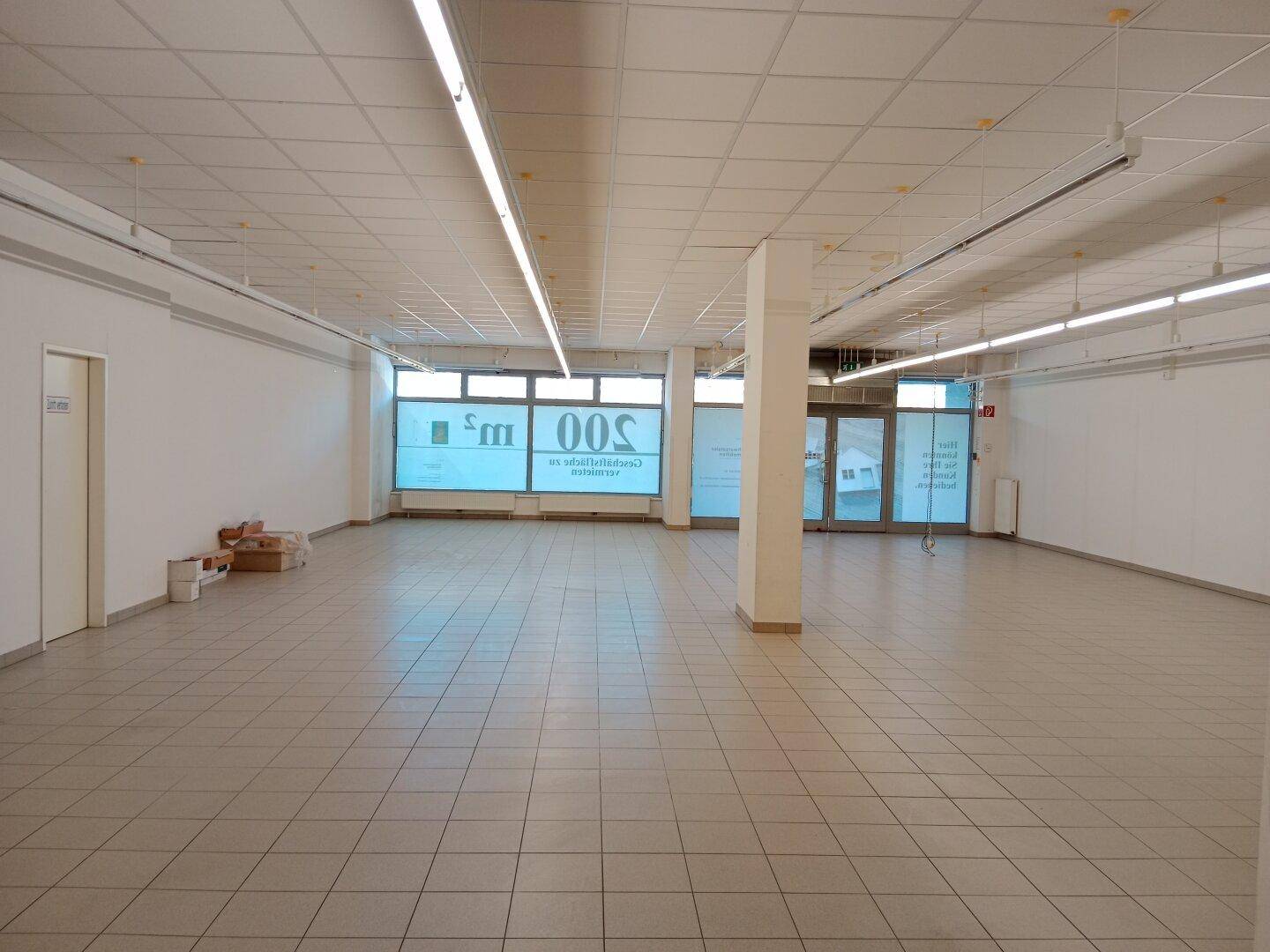 Großer, leerer Verkaufsraum mit Fliesenboden, Rasterdecke und großen Schaufenstern, die viel Tageslicht hereinlassen.