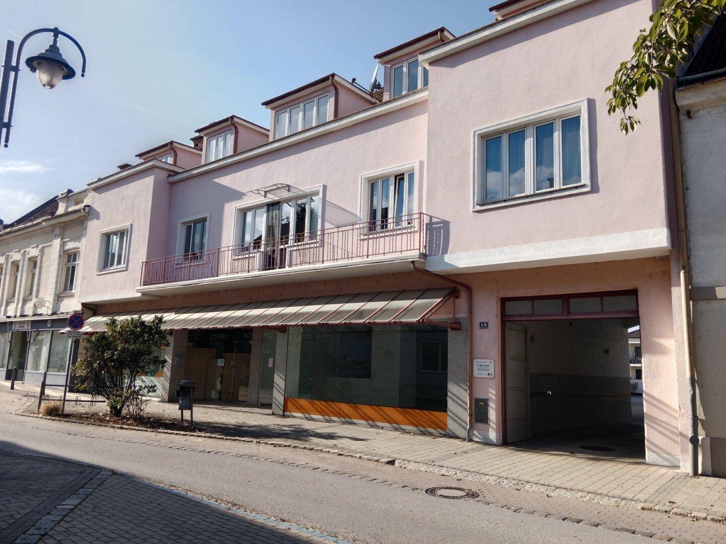 Rosa Gebäude mit Geschäftsflächen im Erdgeschoss und Wohnungen darüber, an einer Straße gelegen.