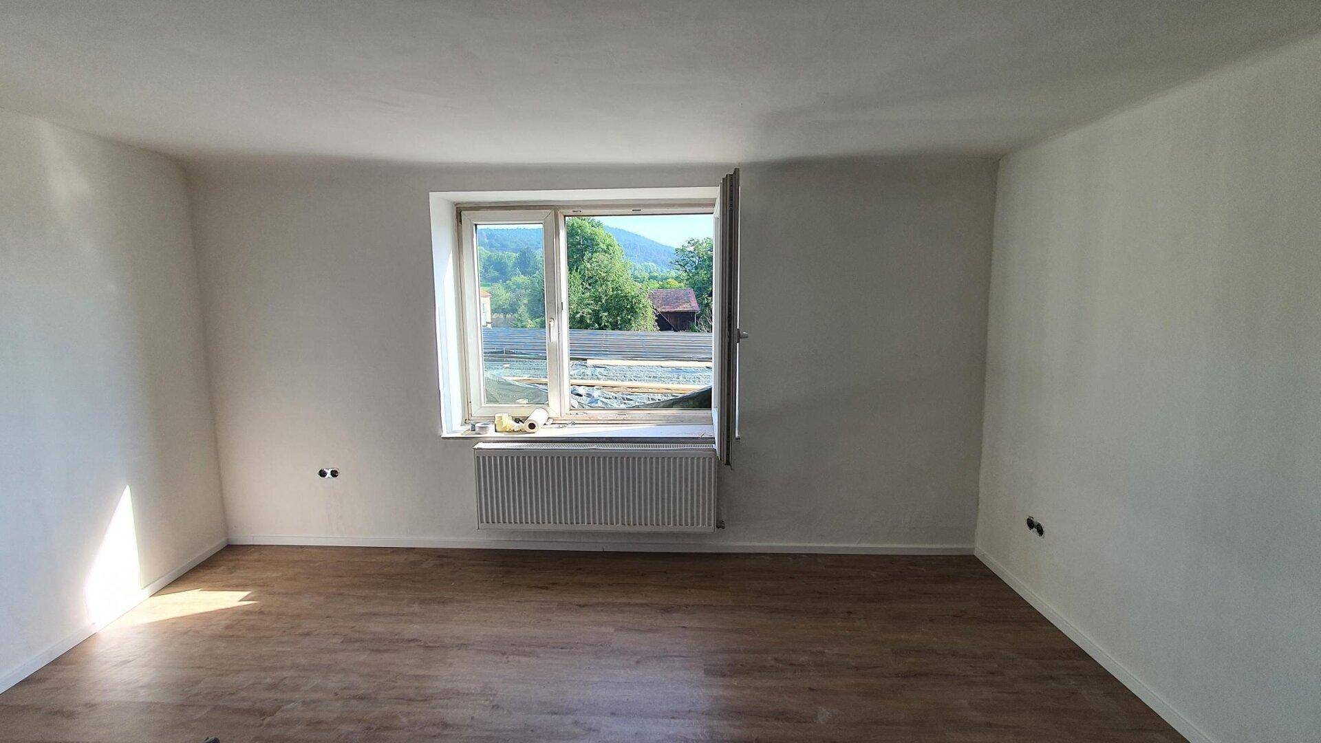 Heller Raum mit Laminatboden und weiß gestrichenen Wänden, mit Fensterblick ins Grüne.