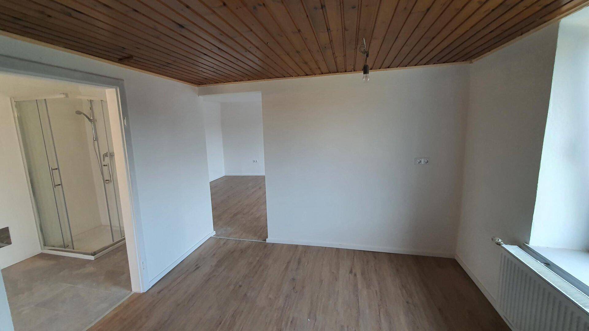 Heller Raum mit Holzdielenboden und Holzdecke, angrenzend an ein Badezimmer mit Duschkabine.