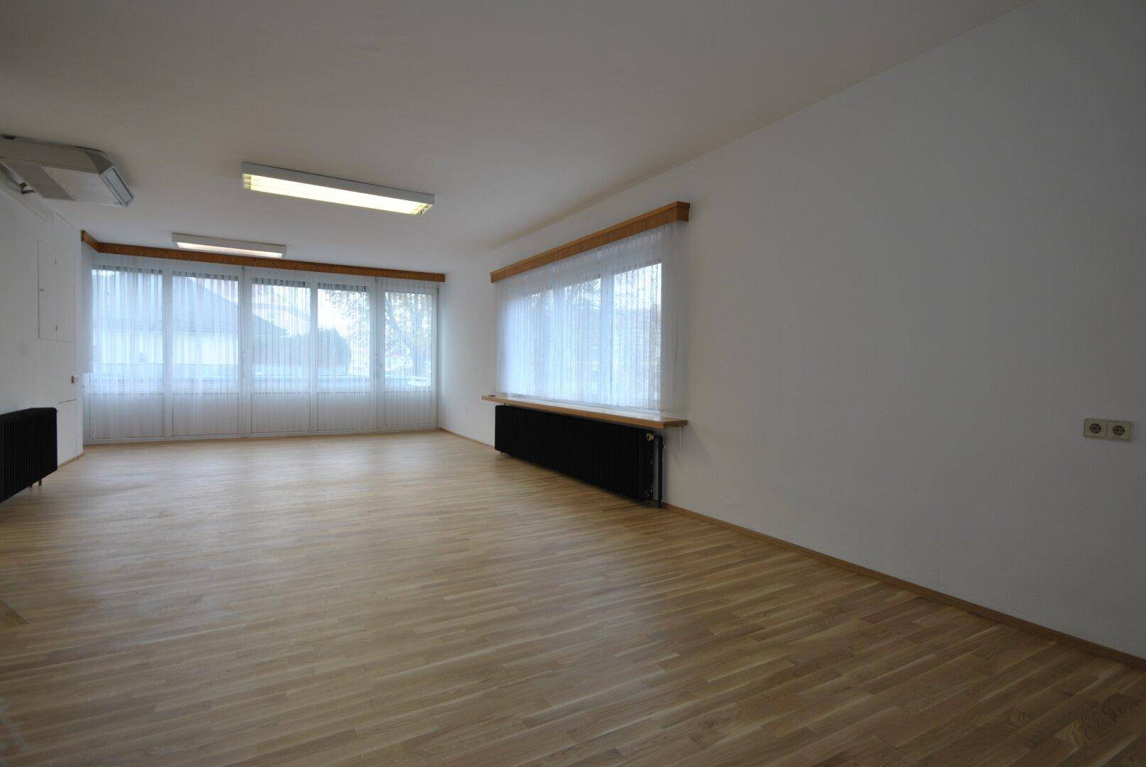 Heller, leerer Raum mit Holzparkettboden, großen Fenstern und Heizkörpern unter den Fenstern.
