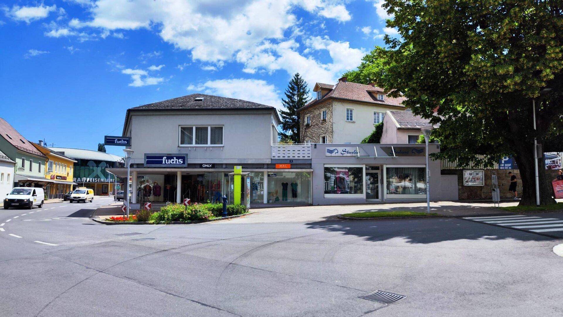 Gebäude mit Geschäftsflächen im Erdgeschoss an einer Straßenecke unter blauem Himmel und Bäumen.