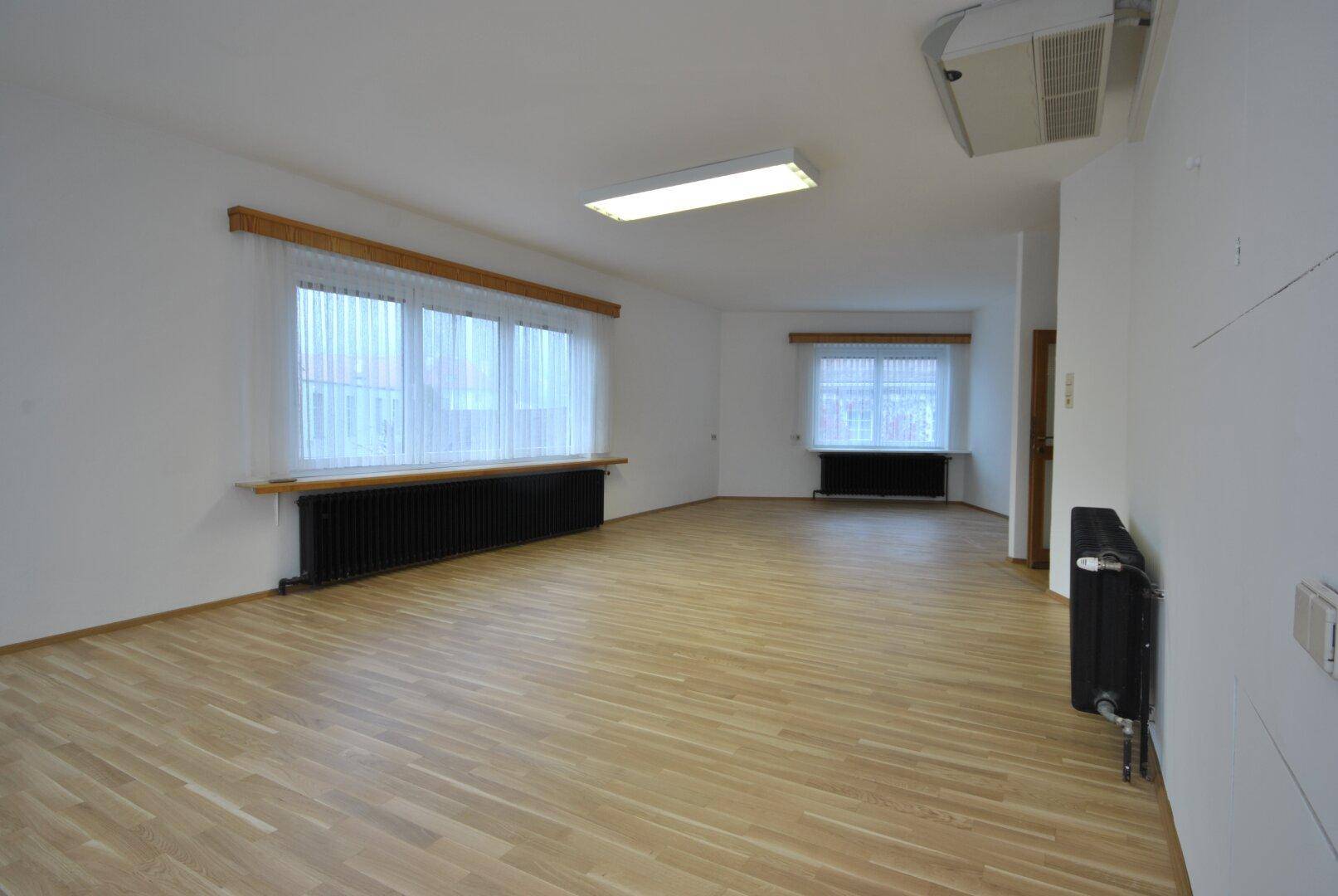 Geräumiger, heller Raum mit Holzboden, großen Fenstern und Deckenbeleuchtung, ideal für Büronutzung.