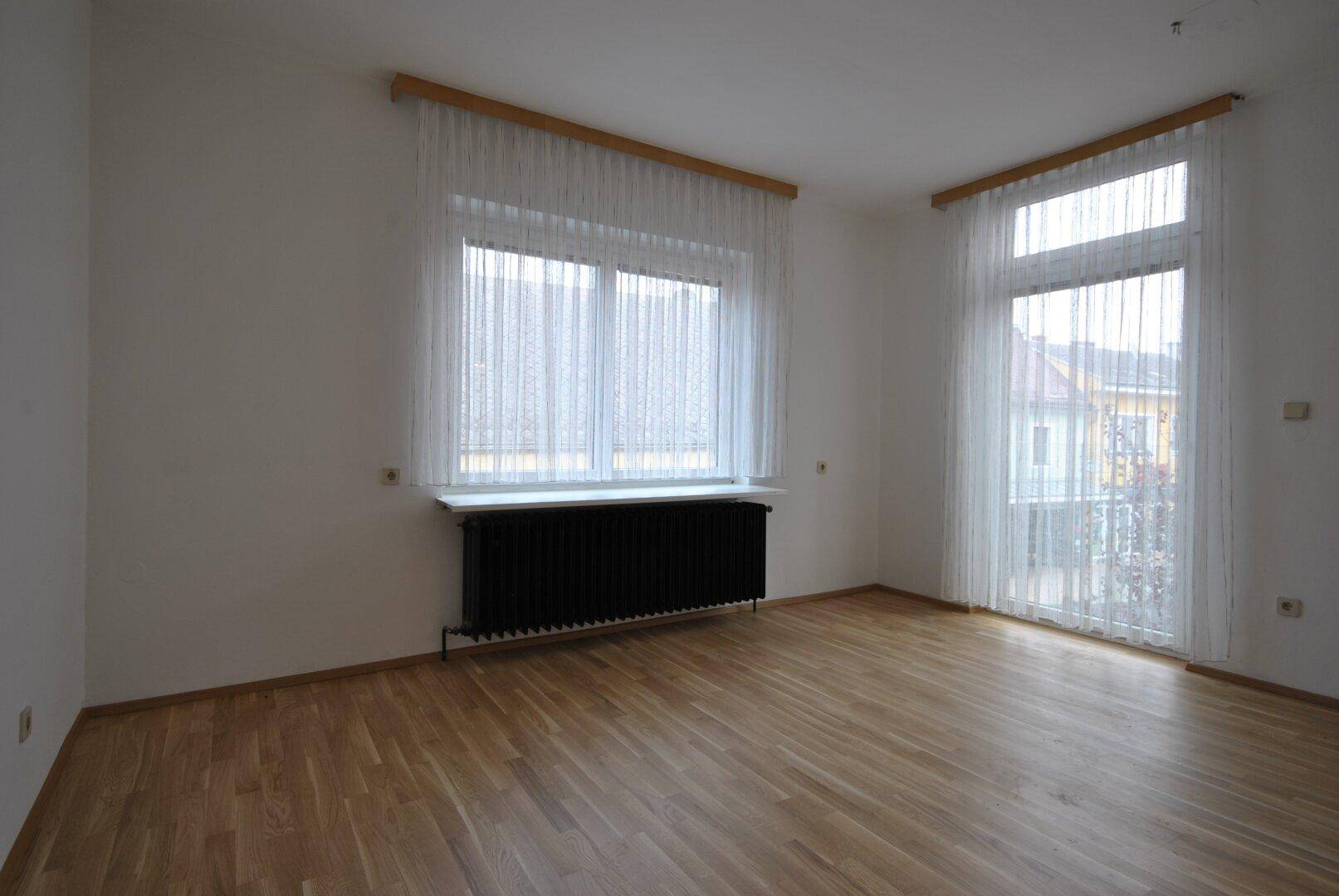 Heller Raum mit Holzboden, großen Fenstern und einem Heizkörper unter den weißen Vorhängen.