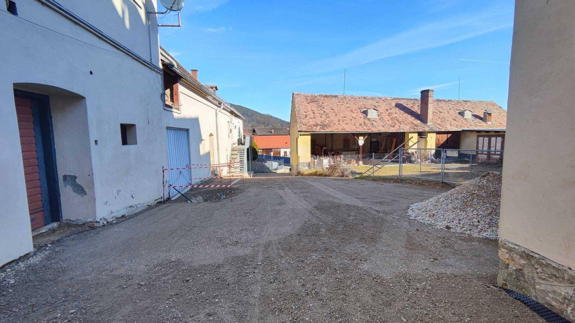 Blick über einen Schotterplatz zwischen mehreren Gebäuden mit Ziegeldächern unter blauem Himmel.