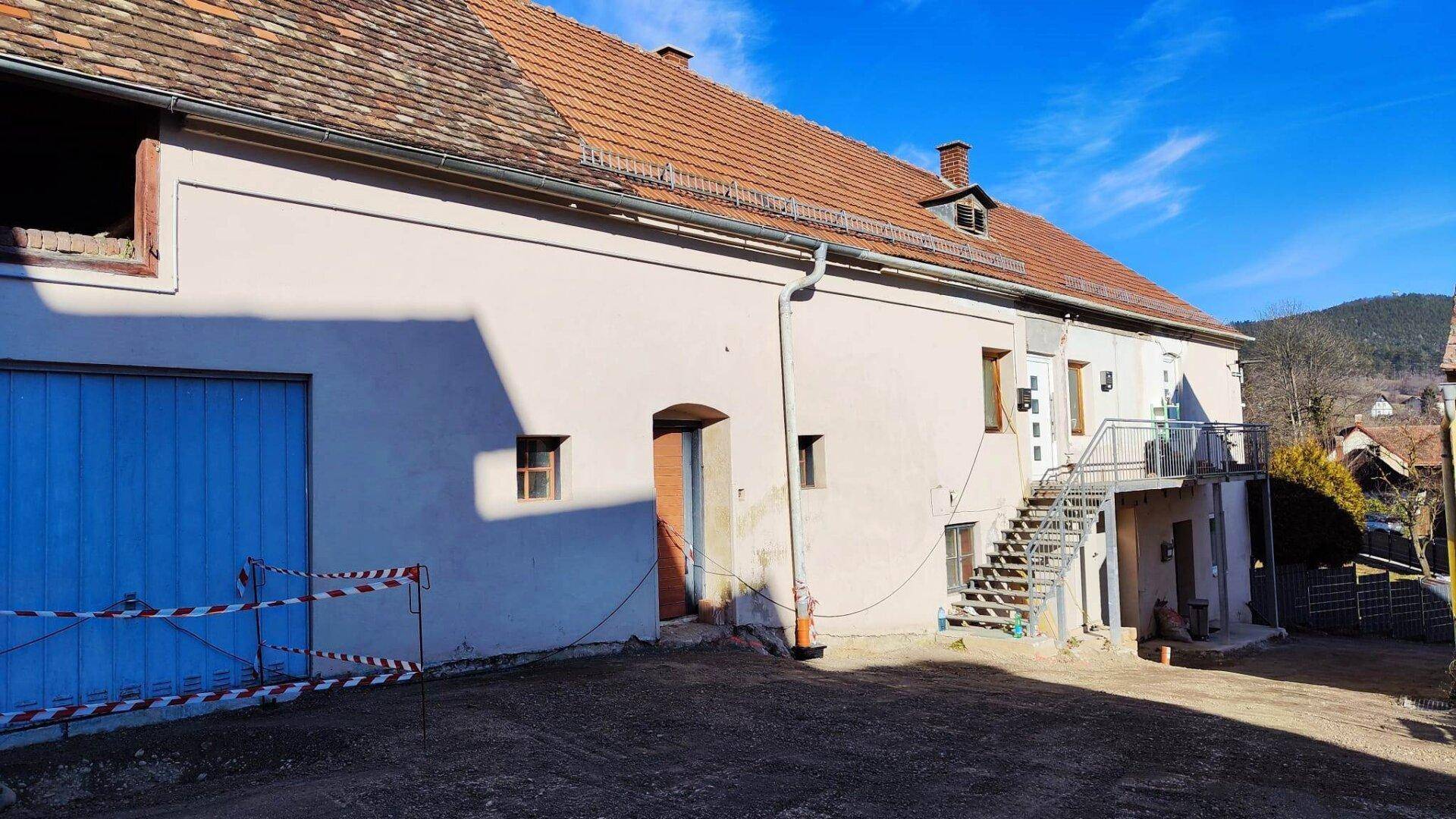 Ansicht eines Gebäudes mit Ziegeldach, Außentreppe und blauen Garagentoren im ländlichen Raum.