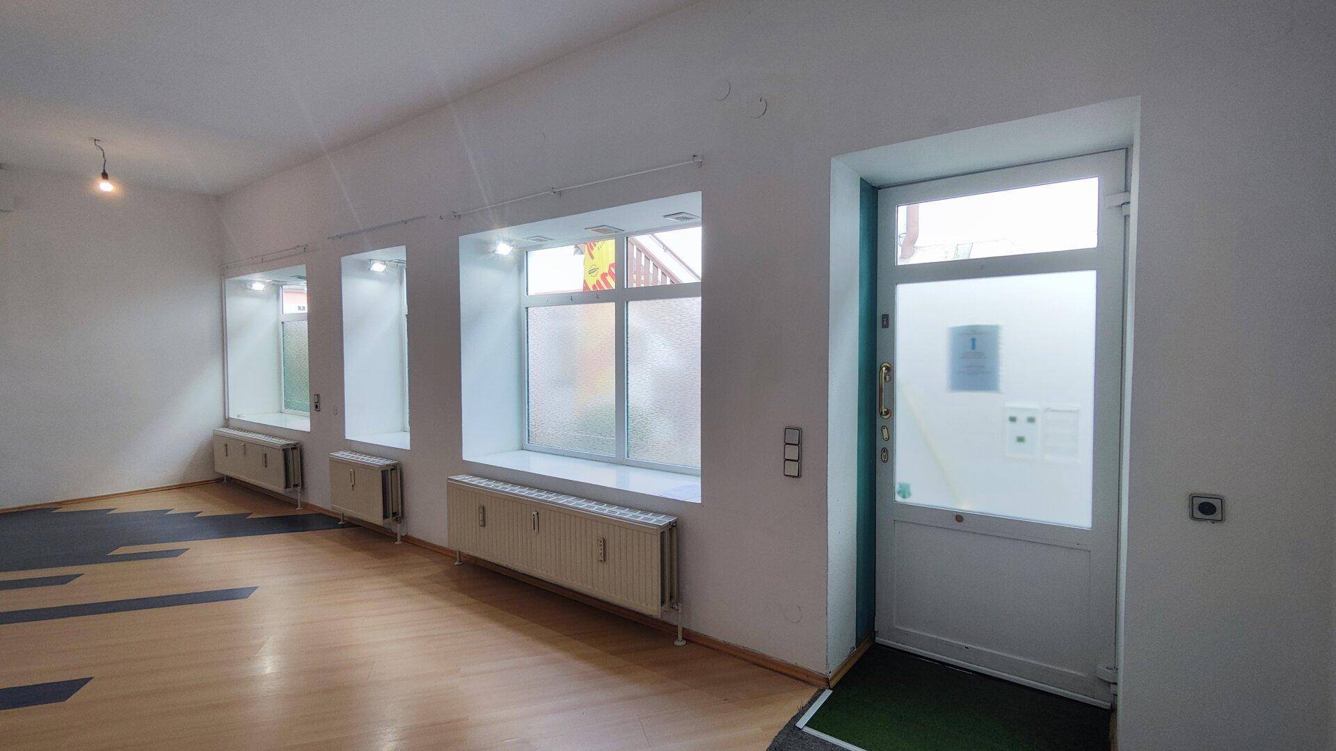 Ein heller, leerer Geschäftsraum mit großen Fenstern, Heizkörpern und einem modernen Laminatboden.