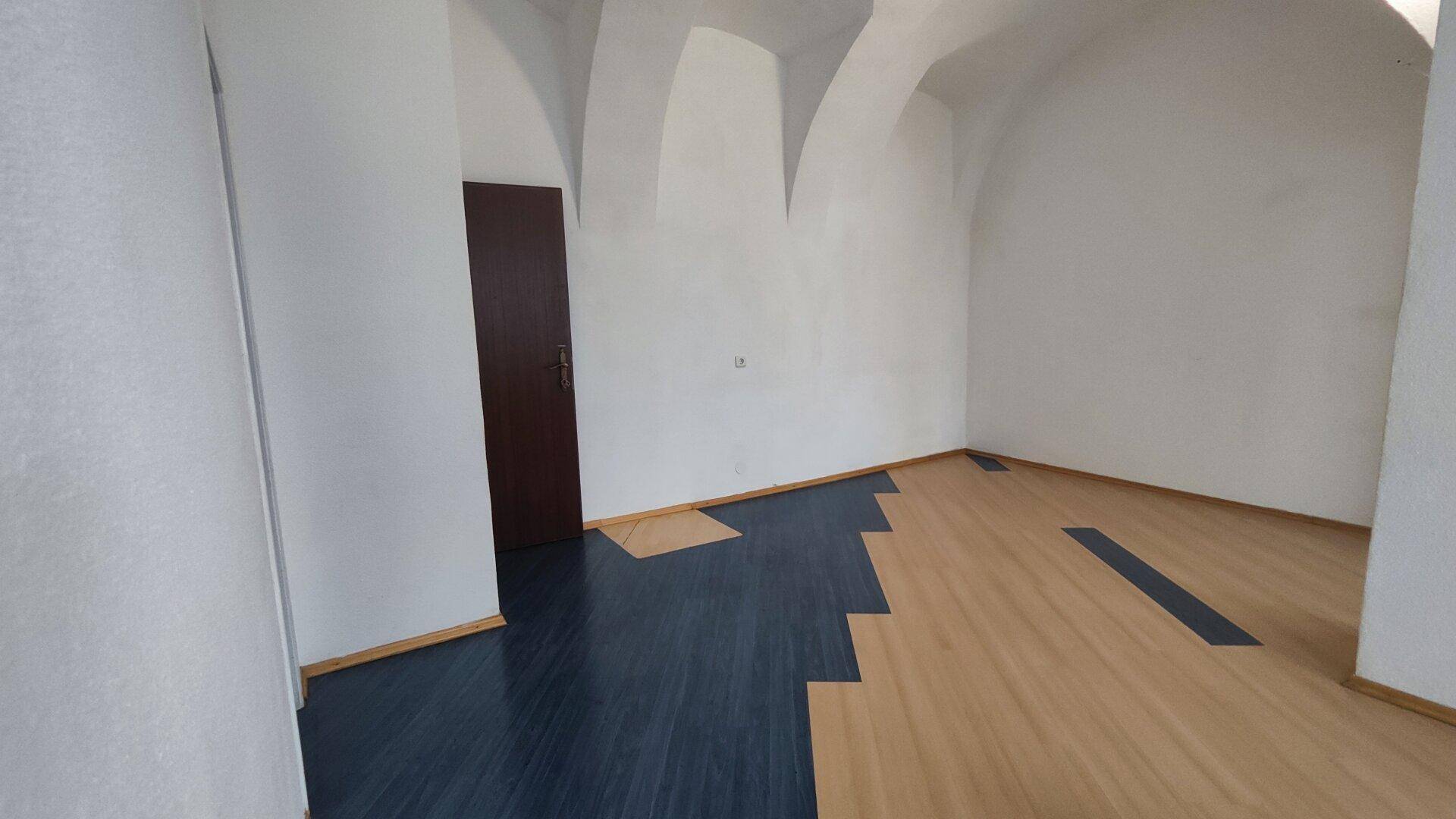 Ein leerer Raum mit gewölbter Decke, einer Holztür und einem auffälligen zweifarbigen Bodenbelag.