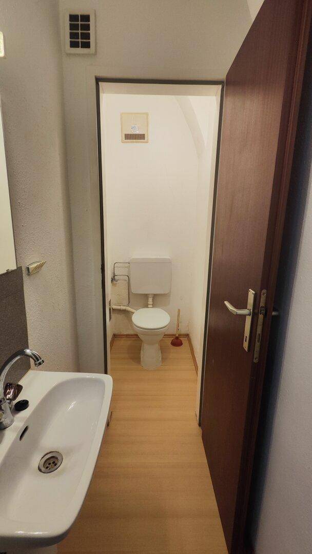 Ein schlichtes Badezimmer mit Waschbecken und Toilette, durch eine offene Tür sichtbar.