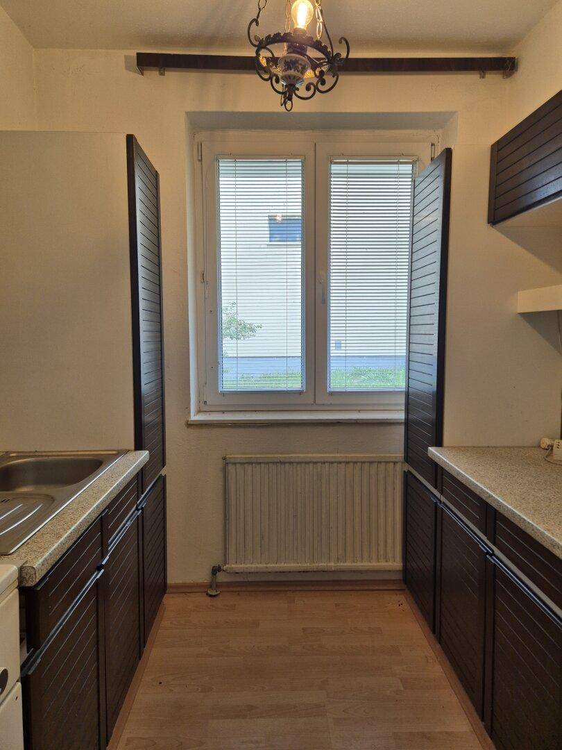 Schmale Küche mit dunklen Holzschränken, Spüle und einem Fenster mit Jalousien.
