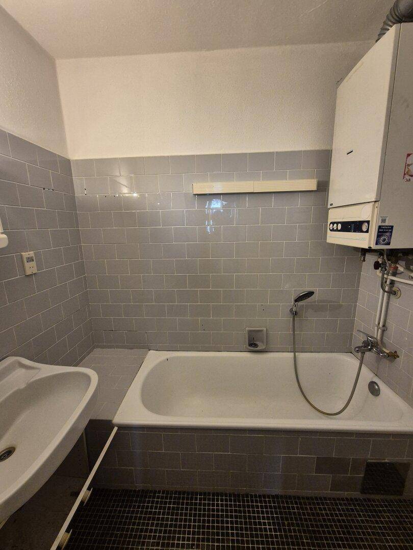 Badezimmer mit grauen Fliesen, Badewanne, Waschbecken und einem Warmwasserboiler.