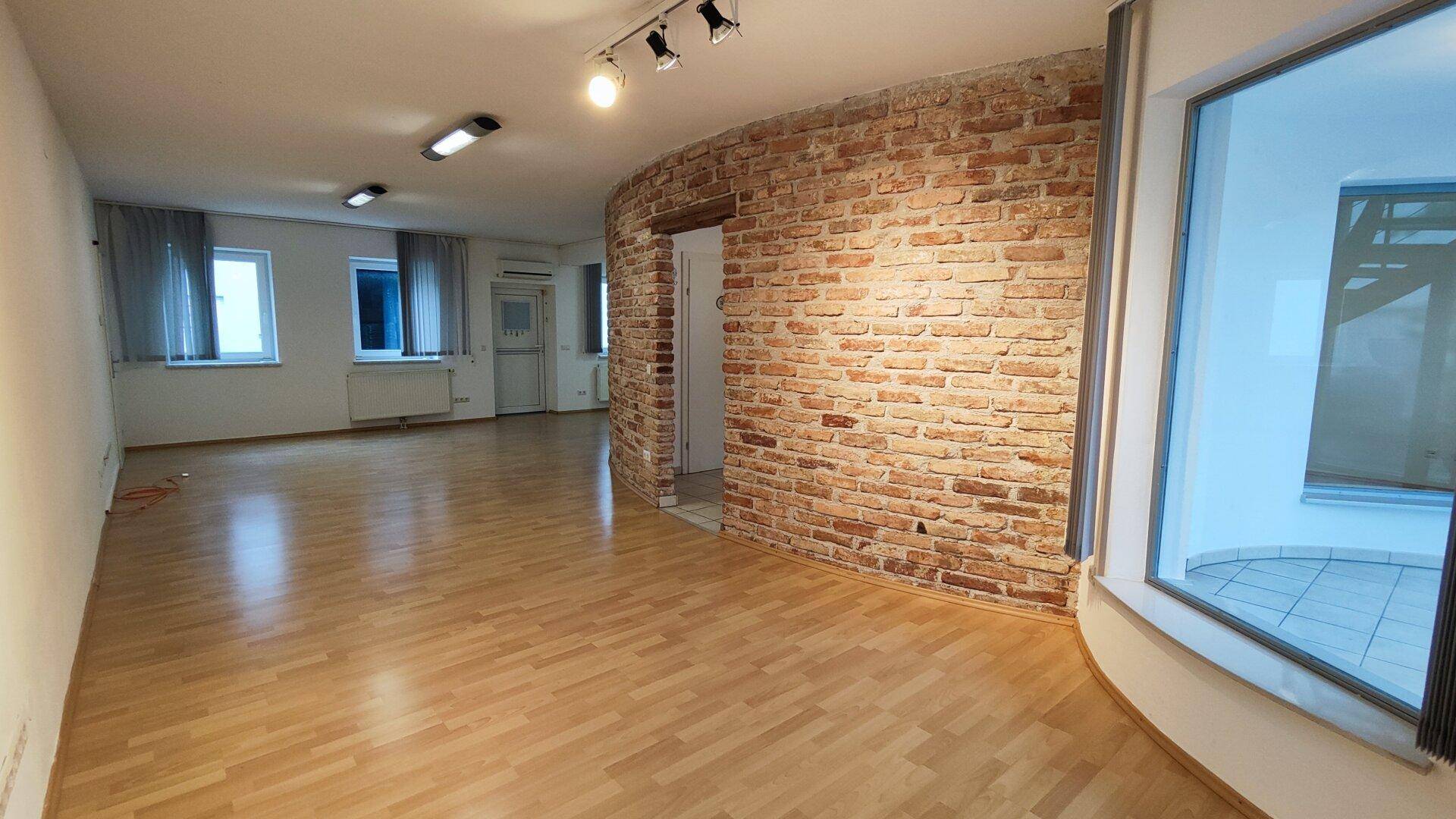 Geräumiger Büroraum mit Holzboden, einer geschwungenen Ziegelwand und großen Fenstern, die den Raum erhellen.