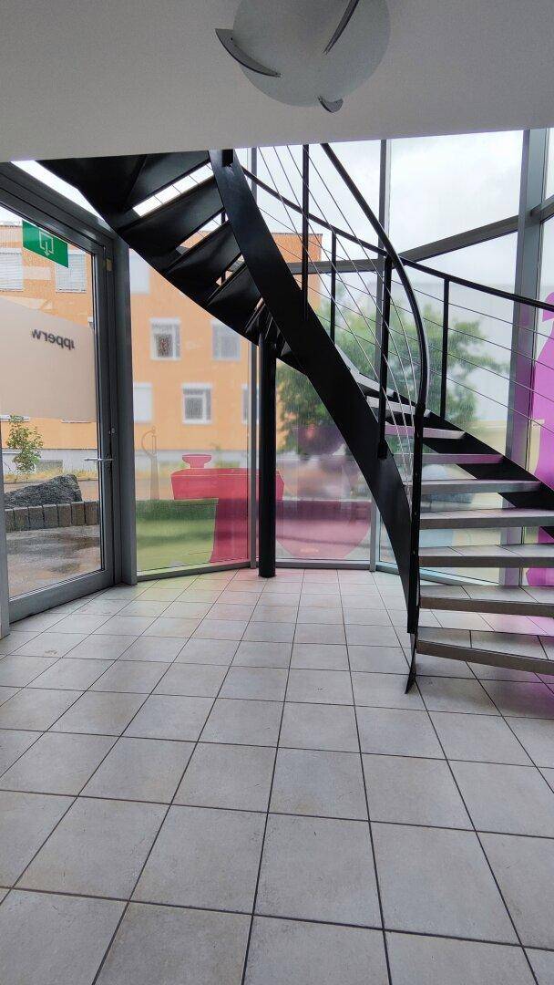 Heller Eingangsbereich mit einer eleganten Wendeltreppe aus Metall und großen Fenstern, die viel Tageslicht hereinlassen.