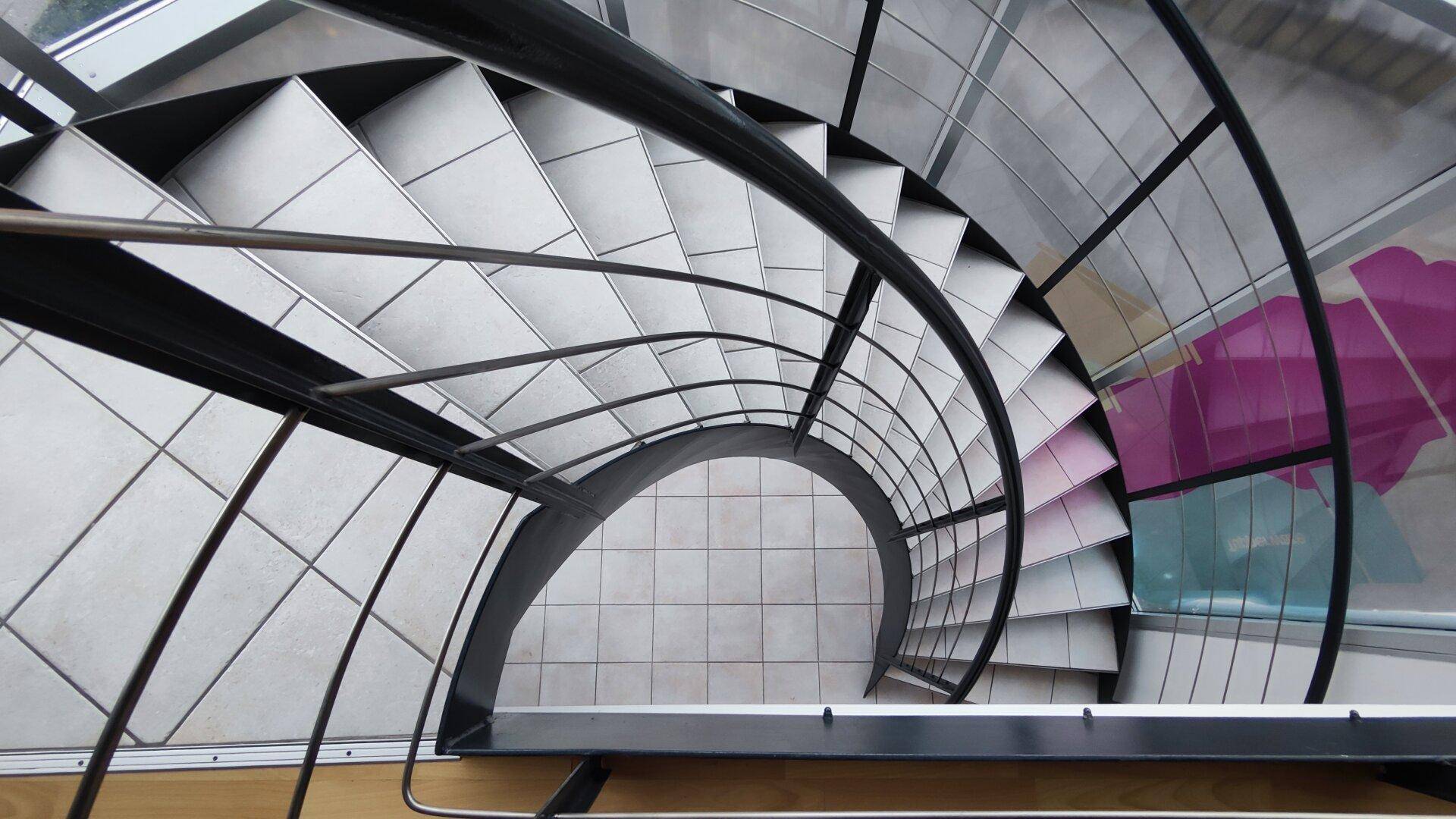 Blick von oben auf eine moderne Wendeltreppe mit Metallgeländer und Fliesenstufen, die nach unten führt.