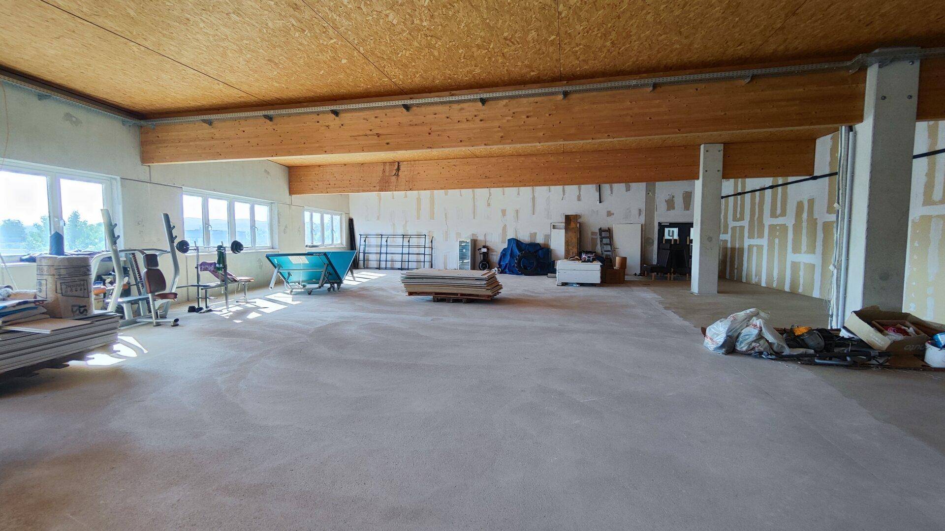 Weitläufiger Gewerberaum mit Holzbalkendecke, Betonboden und viel Tageslicht durch die Fensterfront.