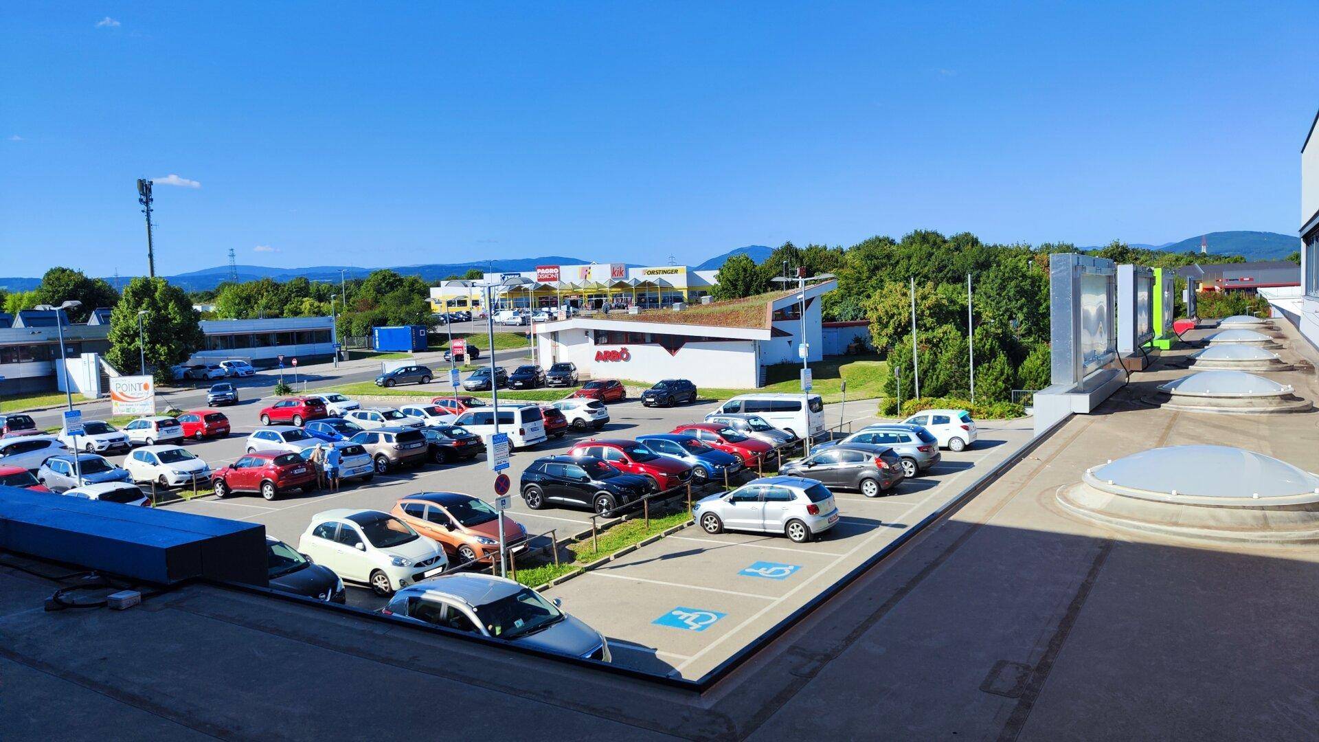Luftaufnahme eines Gewerbegebiets mit großem Parkplatz, begrünten Dächern und Bergen im Hintergrund.
