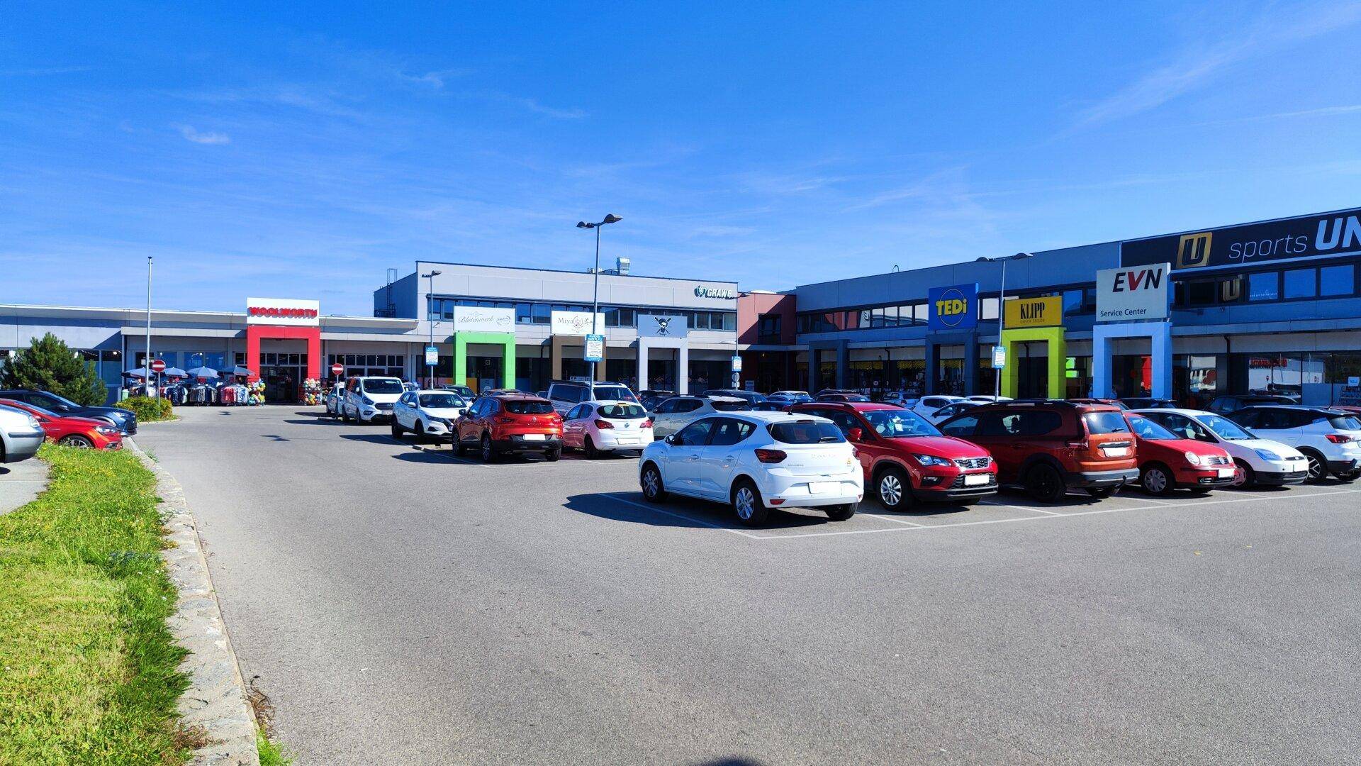 Großer Parkplatz vor einem modernen Einkaufszentrum mit verschiedenen Geschäften unter blauem Himmel.