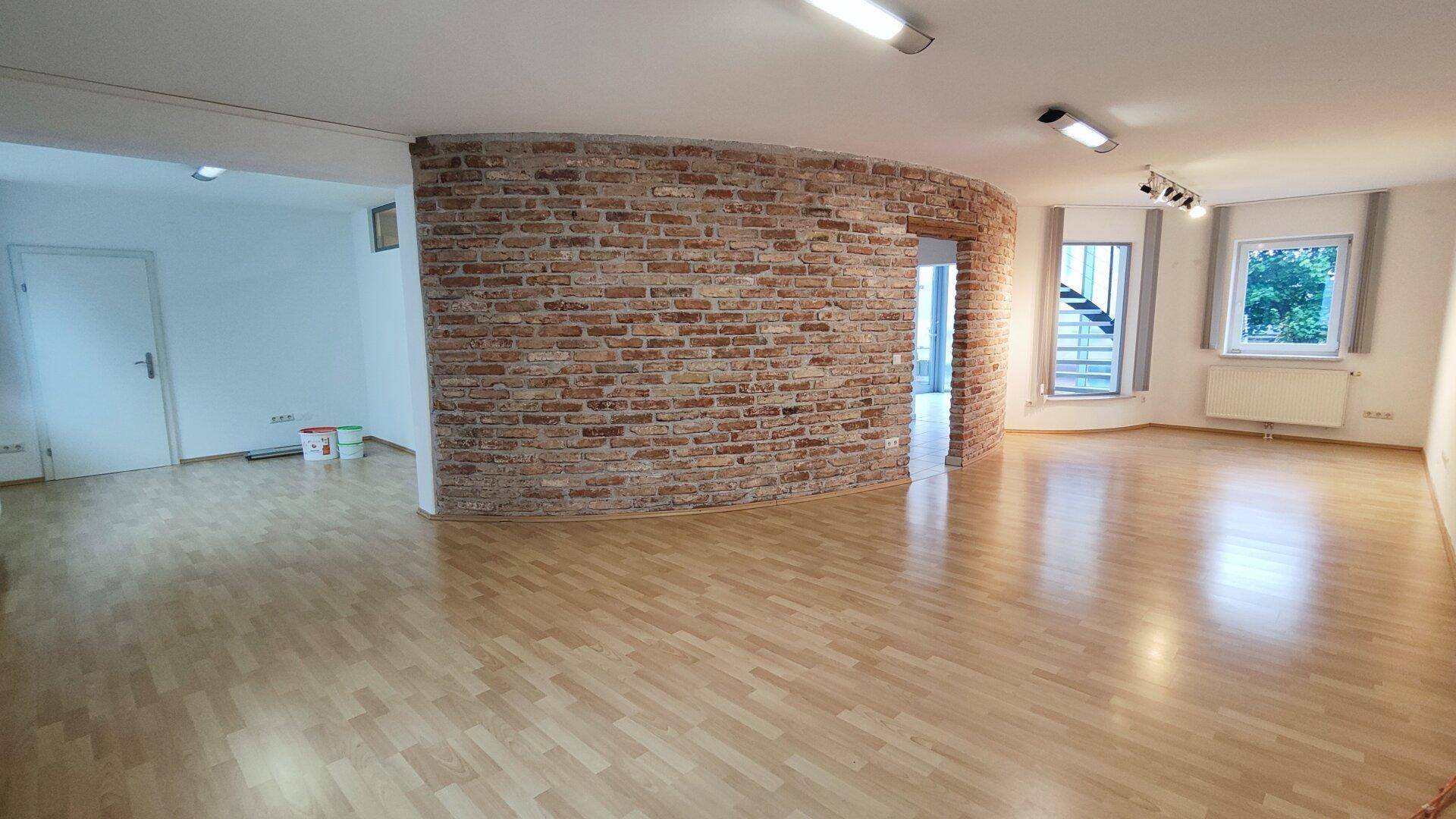 Heller Büroraum mit Holzboden und einer markanten Ziegelwand, die den Raum in verschiedene Bereiche unterteilt.