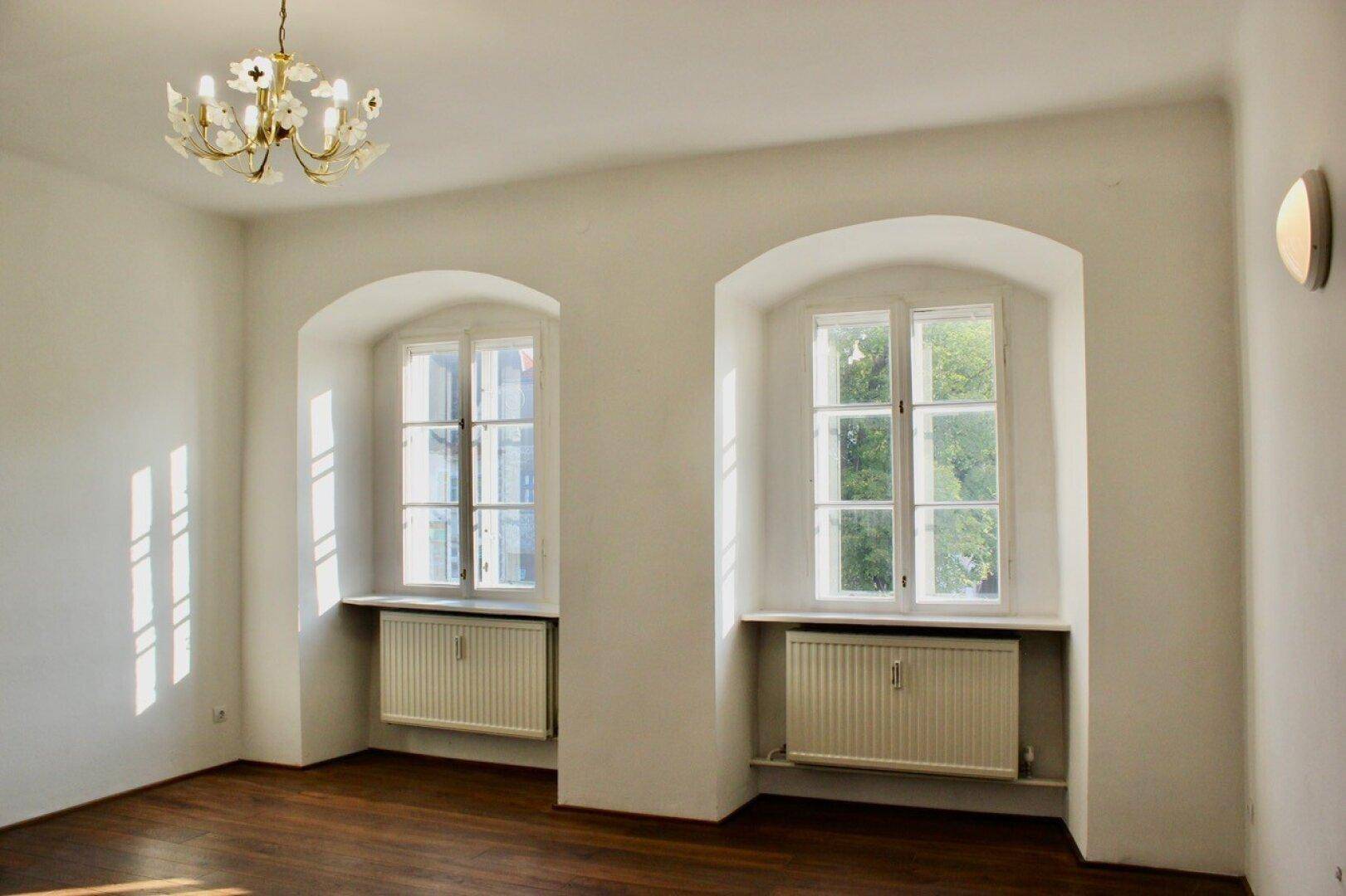 Geräumiger Raum mit zwei großen Fenstern, Kronleuchter und dunklem Holzboden, bietet viel Tageslicht.