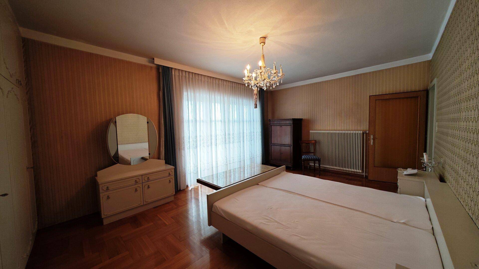 Schlafzimmer mit Doppelbett, Kommode und großen Fenstern, klassisch eingerichtet.