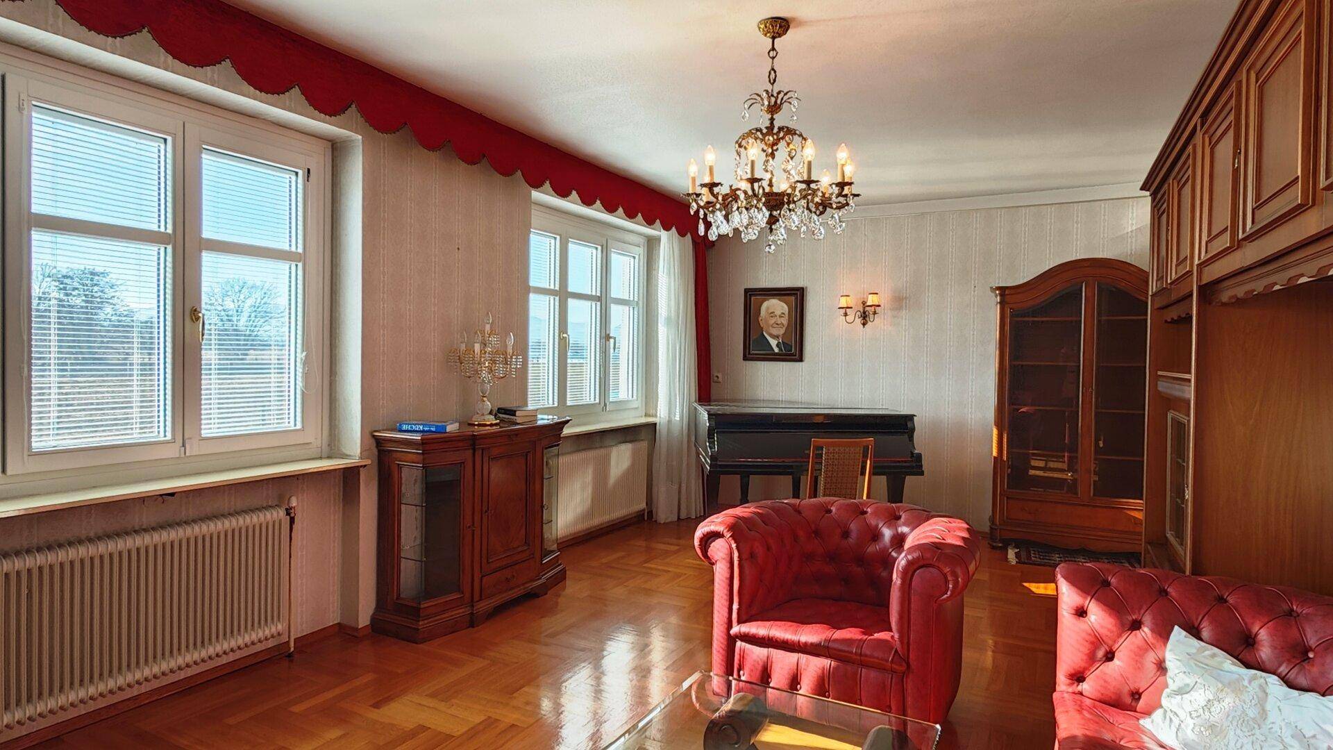 Helles Wohnzimmer mit Flügel, roten Ledersesseln und großen Fenstern.
