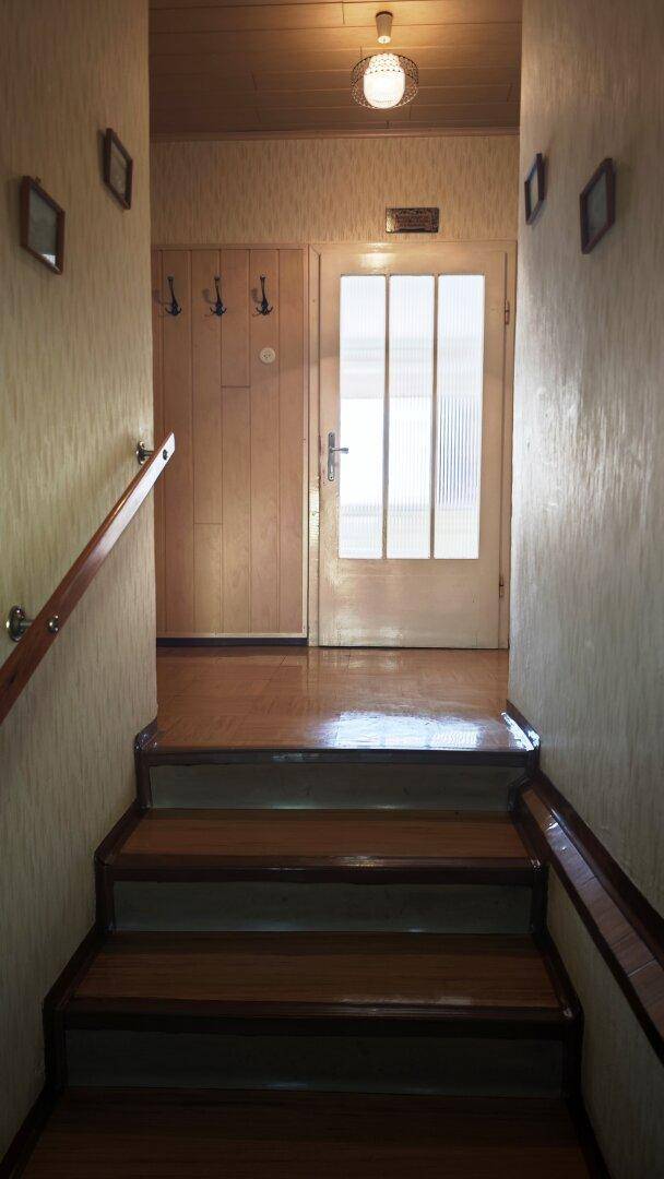 Holztreppe führt zu einem Flur mit gemusterter Tapete und einer Tür mit Glaseinsätzen.