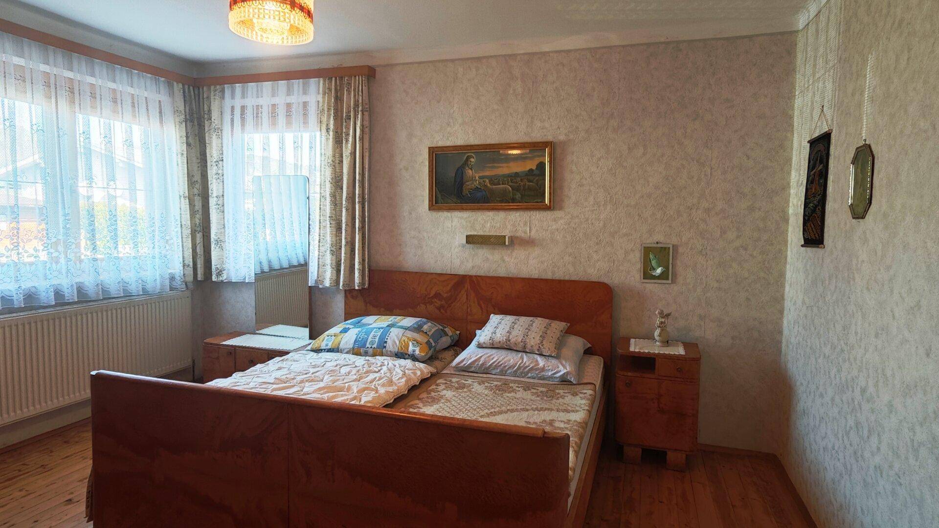 Schlafzimmer mit Doppelbett, Holzmöbeln und gemusterter Tapete, hell durch große Fenster.