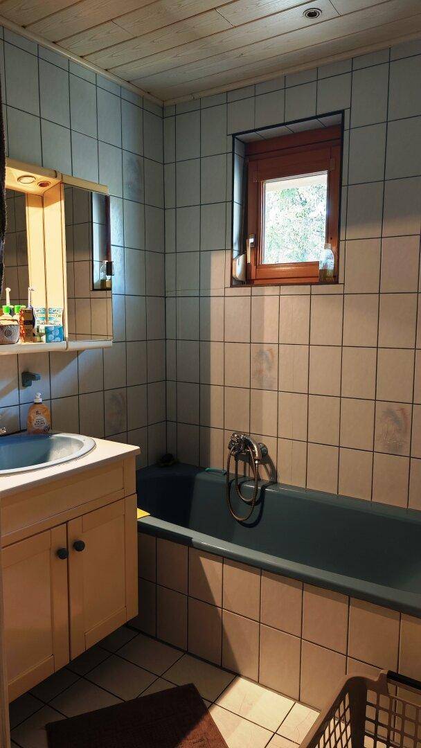 Badezimmer mit Badewanne, Fliesenwänden und Holzdecke, hell durch ein Fenster.