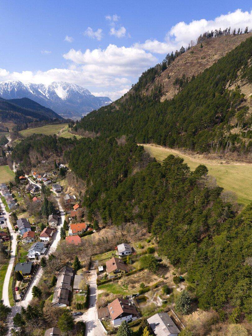 Luftaufnahme eines Dorfes in einem Tal, umgeben von bewaldeten Bergen und schneebedeckten Gipfeln.