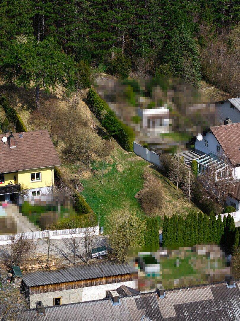 Nähere Luftaufnahme von Wohnhäusern und einem grünen Grundstück am Hang mit Wald im Hintergrund.