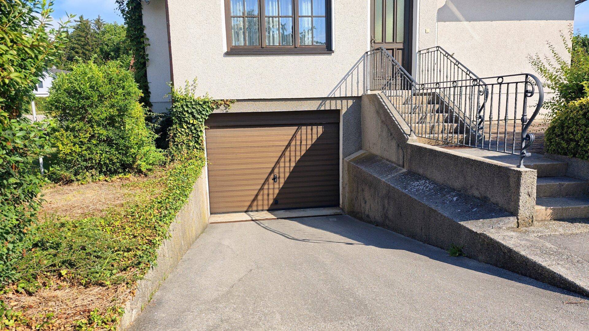 Einfahrt zur Tiefgarage unter dem Haus mit braunem Garagentor und Steintreppe zum Eingang.