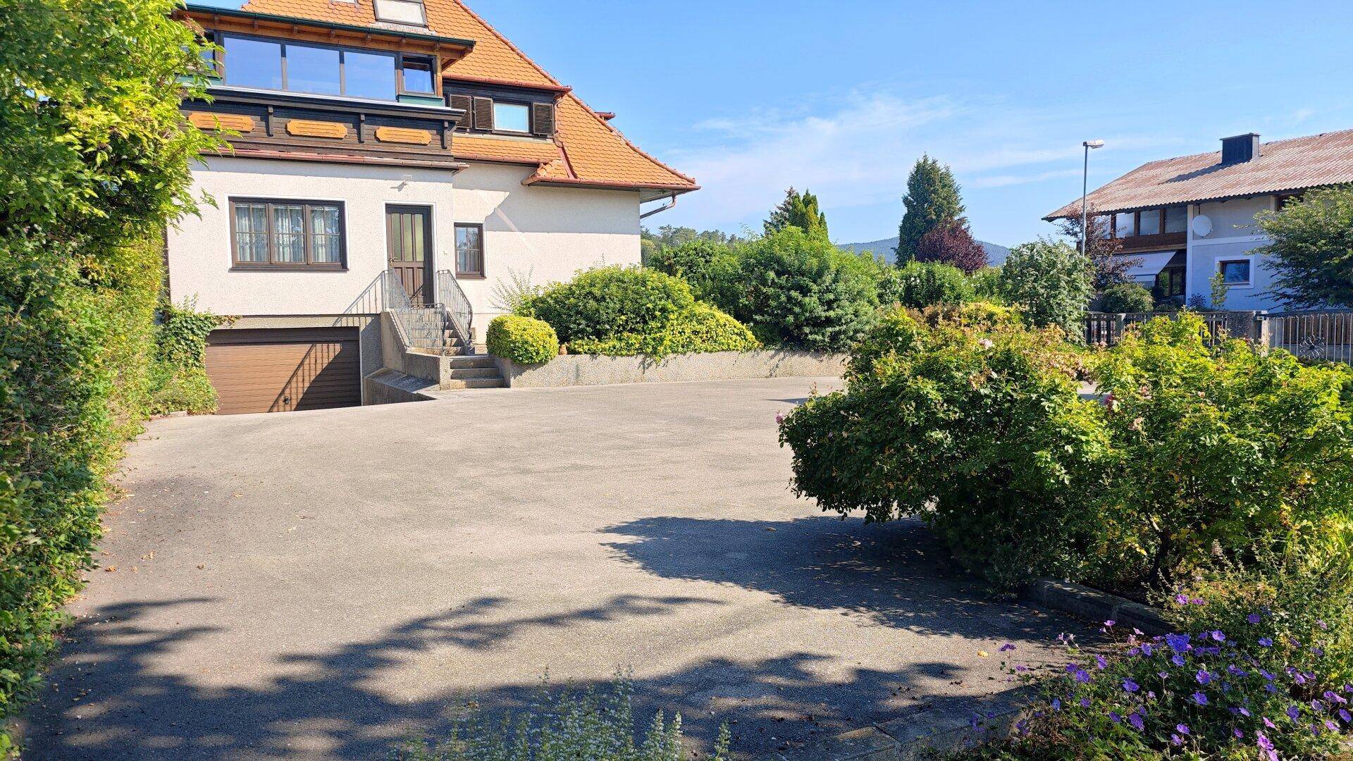 Großzügiger Vorplatz mit viel Parkfläche vor dem Haus, umgeben von grünen Büschen und Bäumen.