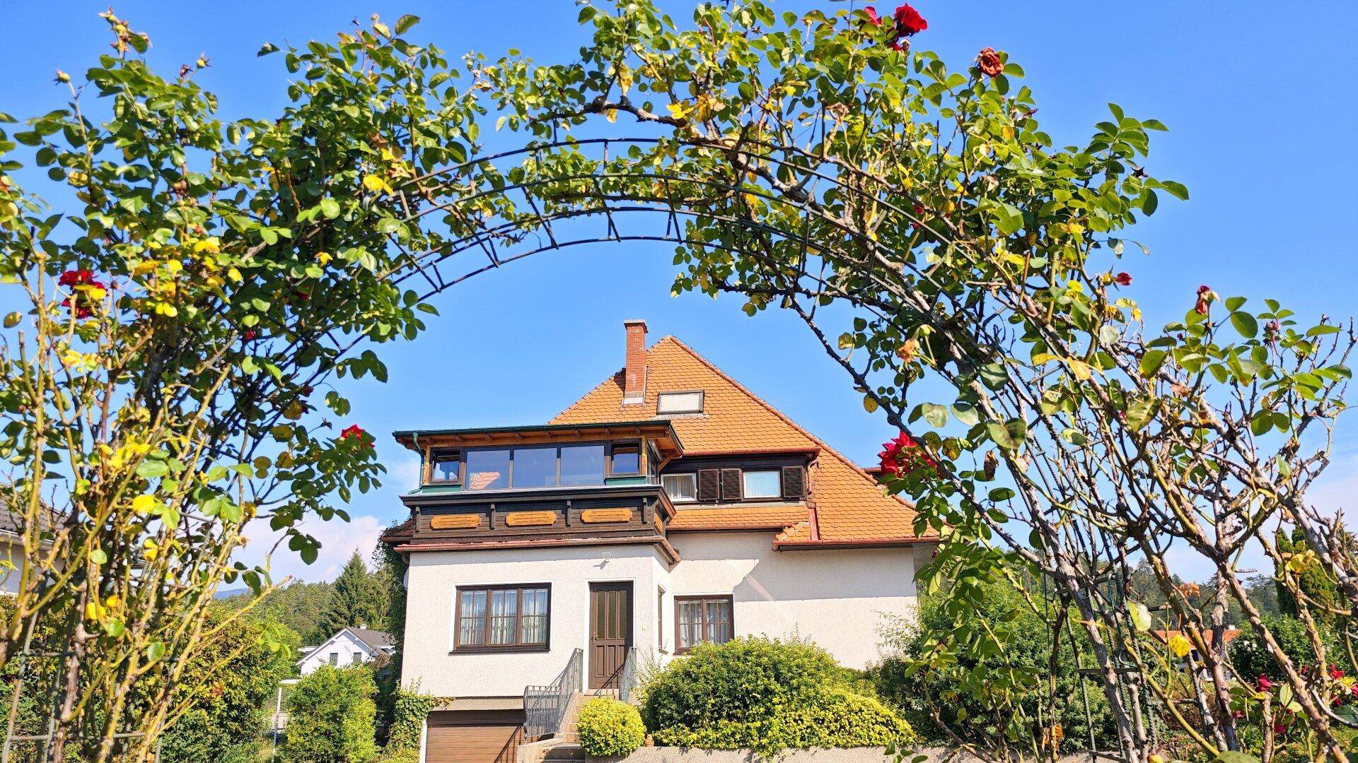 Charmantes Haus mit rotem Ziegeldach, umgeben von einem blühenden Rosenbogen im Vorgarten.