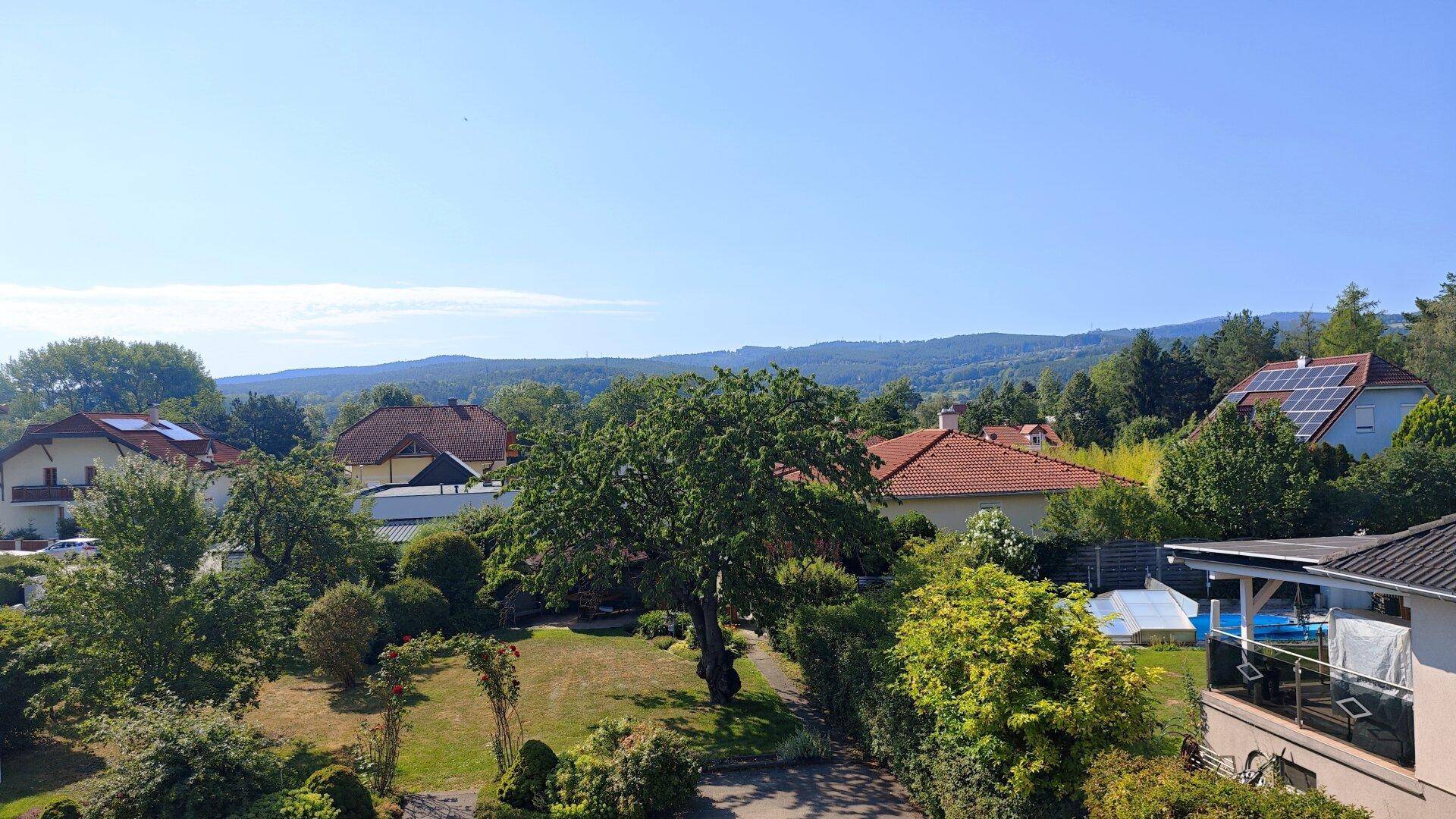 Weitläufiger Blick über die grüne Landschaft mit Häusern und Hügeln unter blauem Himmel.