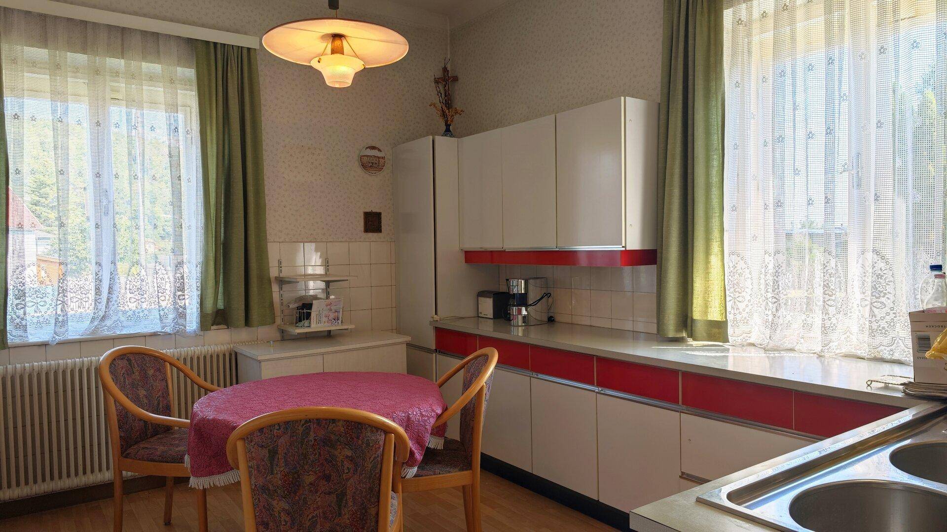 Helle Küche mit weißen und roten Schränken, Esstisch und zwei Fenstern, die viel Licht hereinlassen.