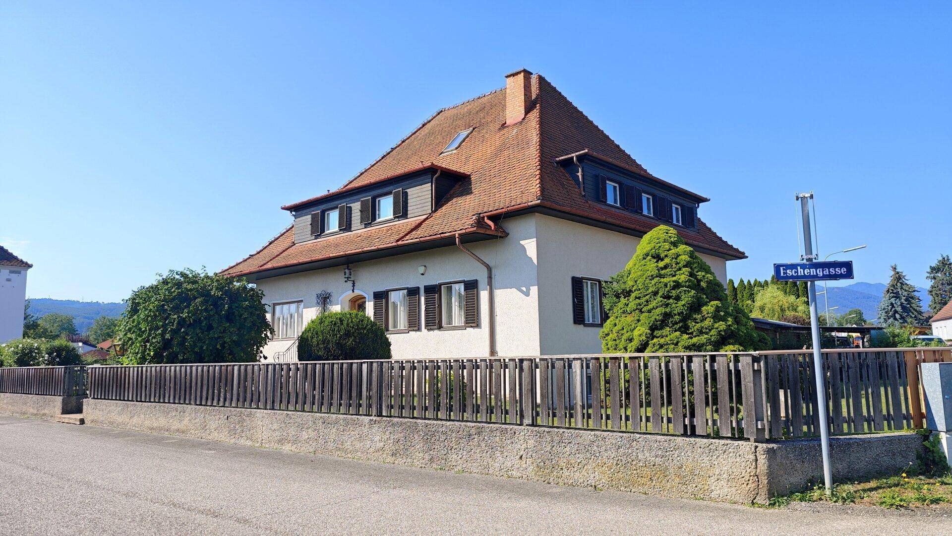 Ansicht des Hauses von der Straße mit Zaun, grünen Büschen und einem Straßenschild.