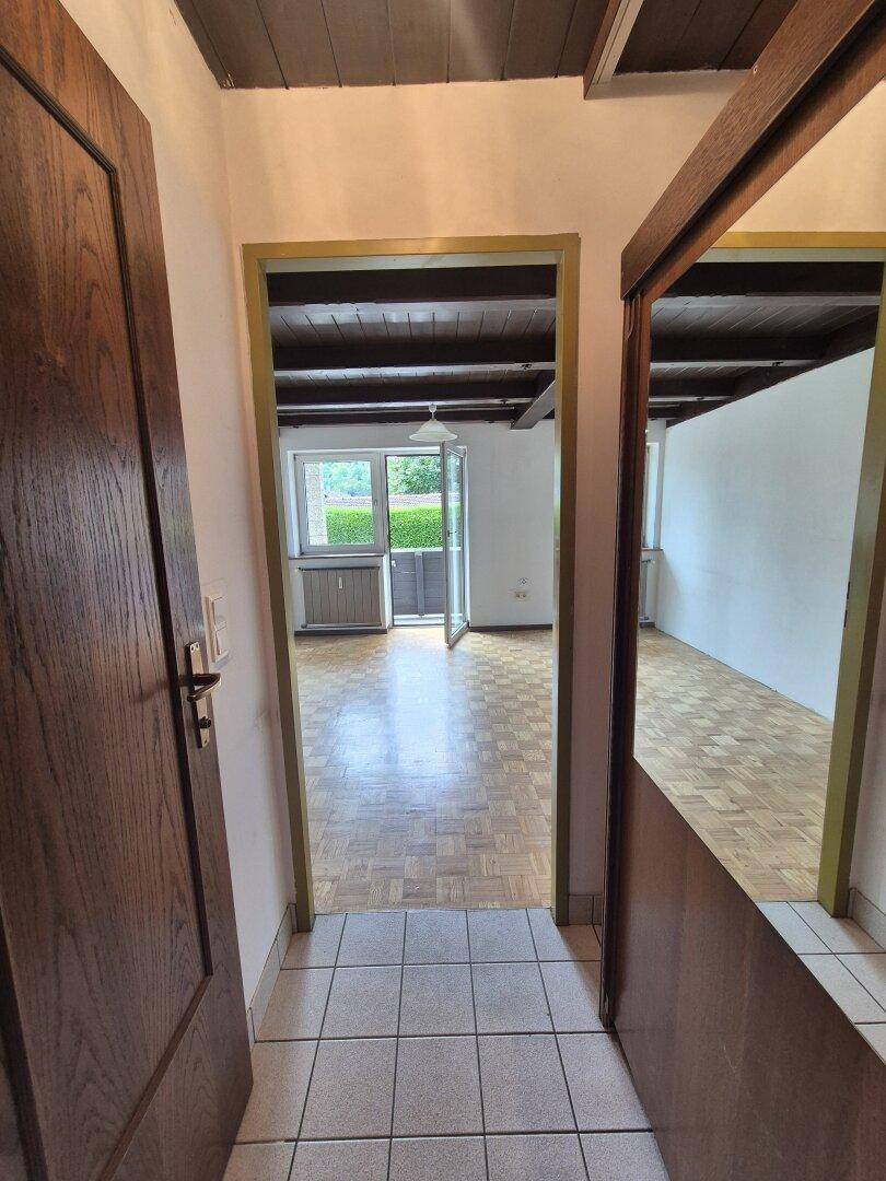 Flur mit Fliesenboden und Holztür, der in einen Raum mit Parkettboden und Holzbalkendecke führt.