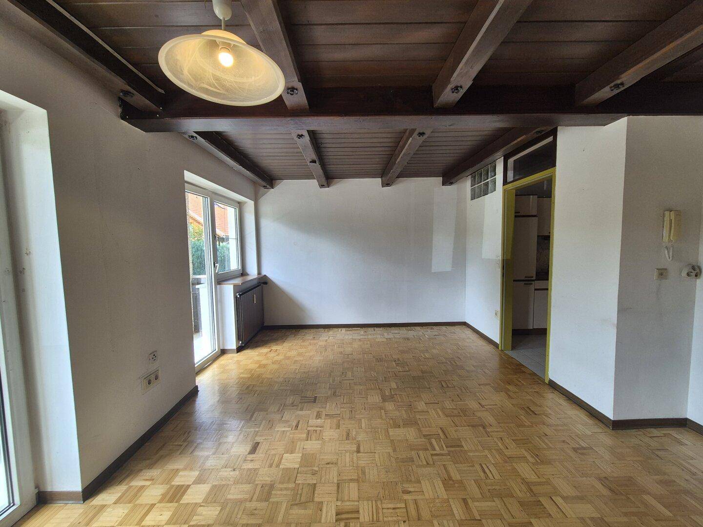 Helles Wohnzimmer mit Parkettboden, Holzbalkendecke und Zugang zum Außenbereich durch große Fenster.