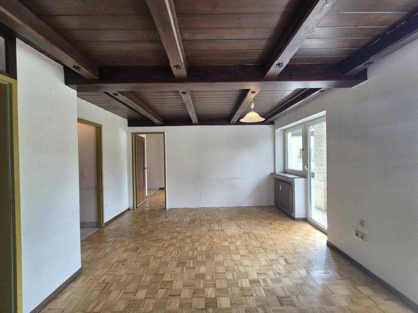 Geräumiges Wohnzimmer mit Parkettboden und sichtbaren Holzbalken an der Decke, gut beleuchtet.