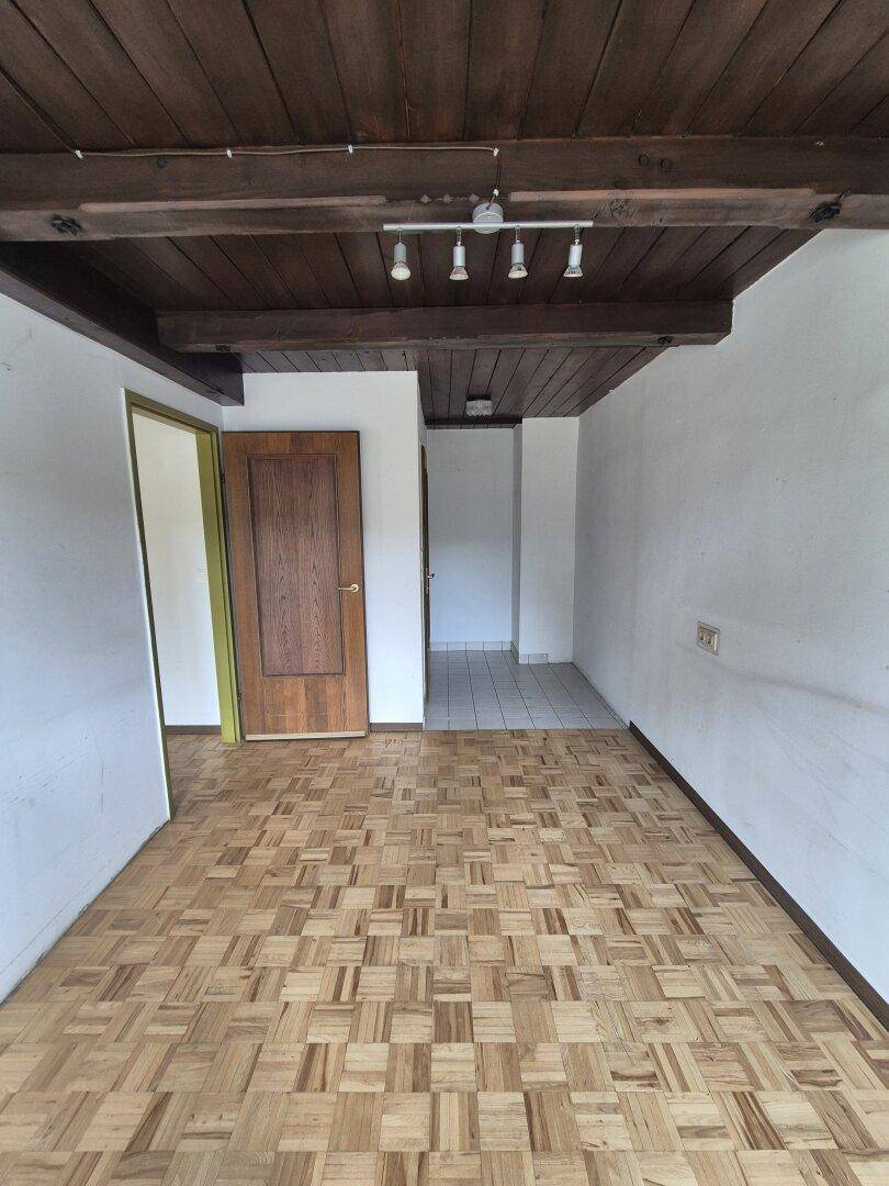 Leerer Raum mit Parkettboden, Holzbalkendecke und Zugang zu einem Flur durch eine Holztür.